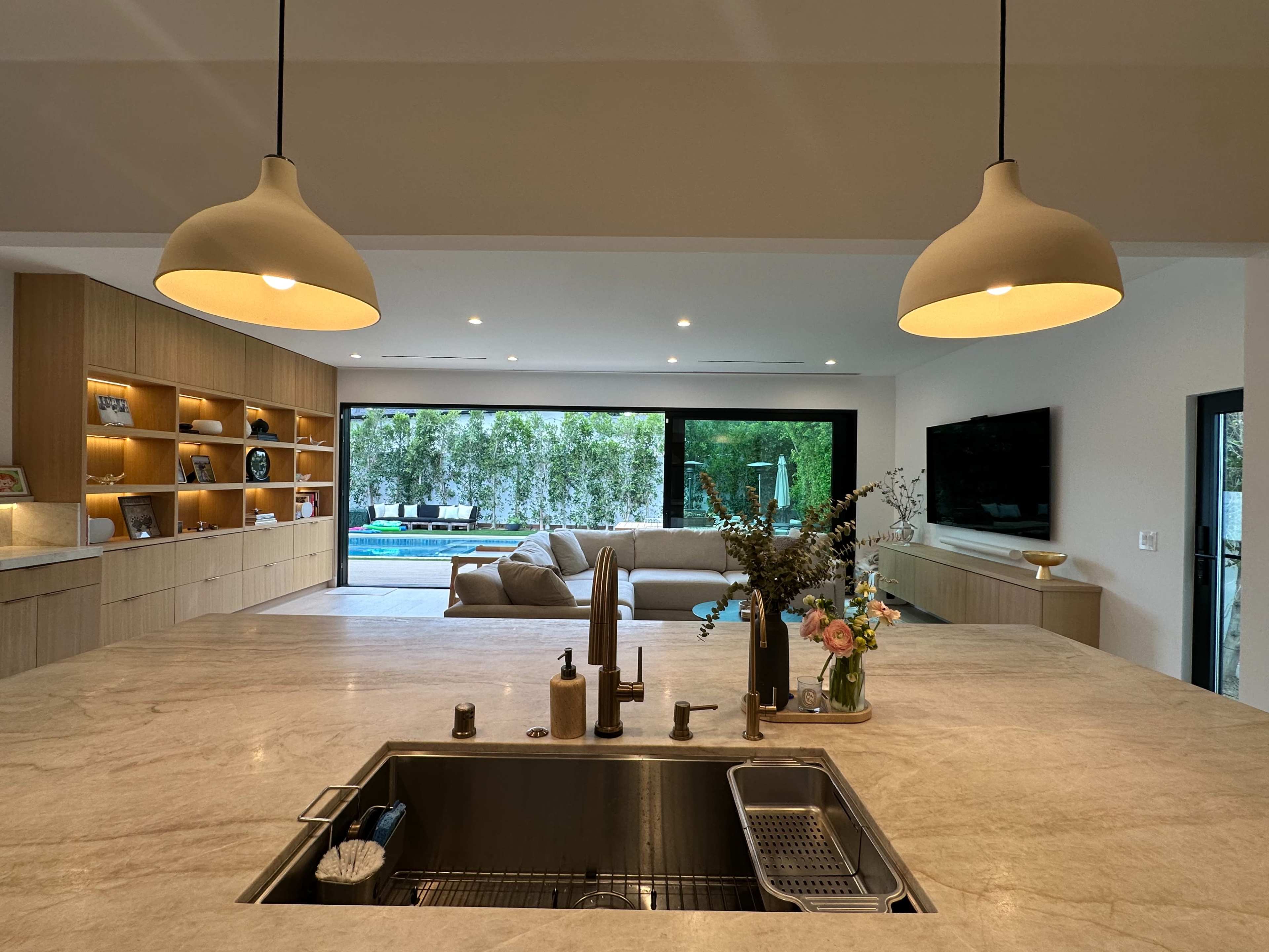 The image depicts a modern kitchen with a marble countertop, sink, and overhead light fixtures, overlooking a living area with large windows that open to a pool and outdoor space.