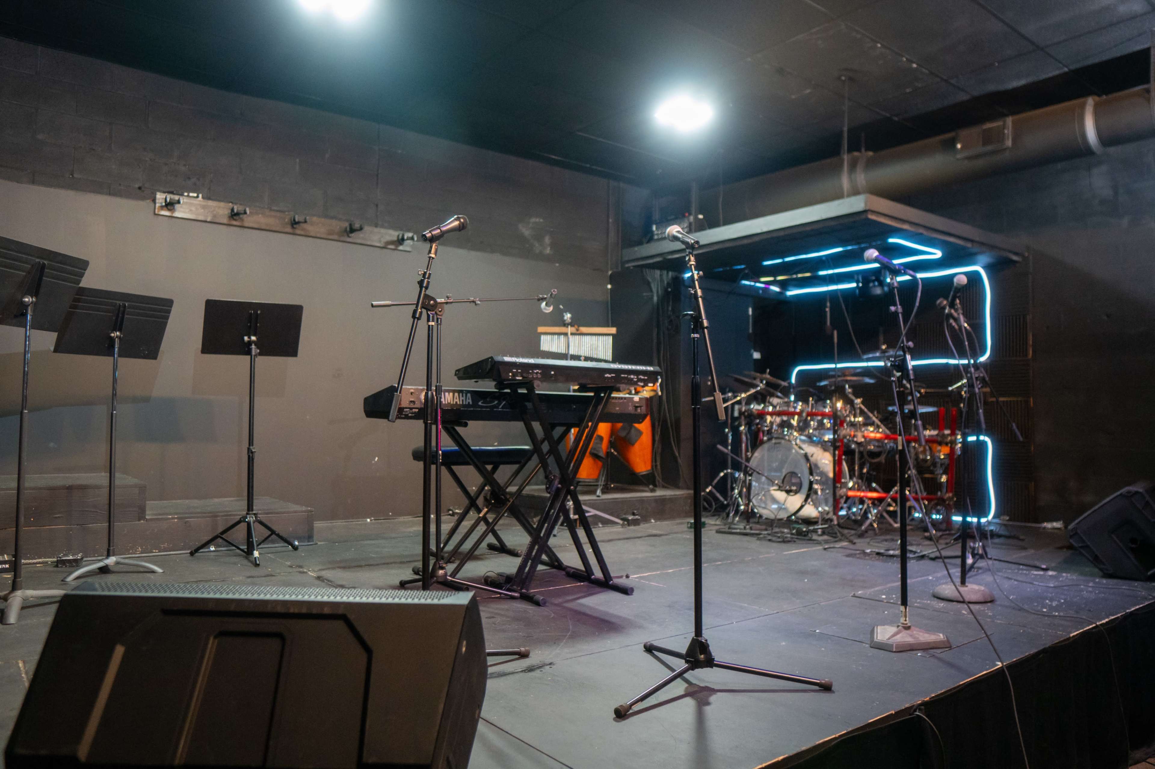 The image shows a performance stage set up with a keyboard, a drum kit, and several microphones positioned on stands.