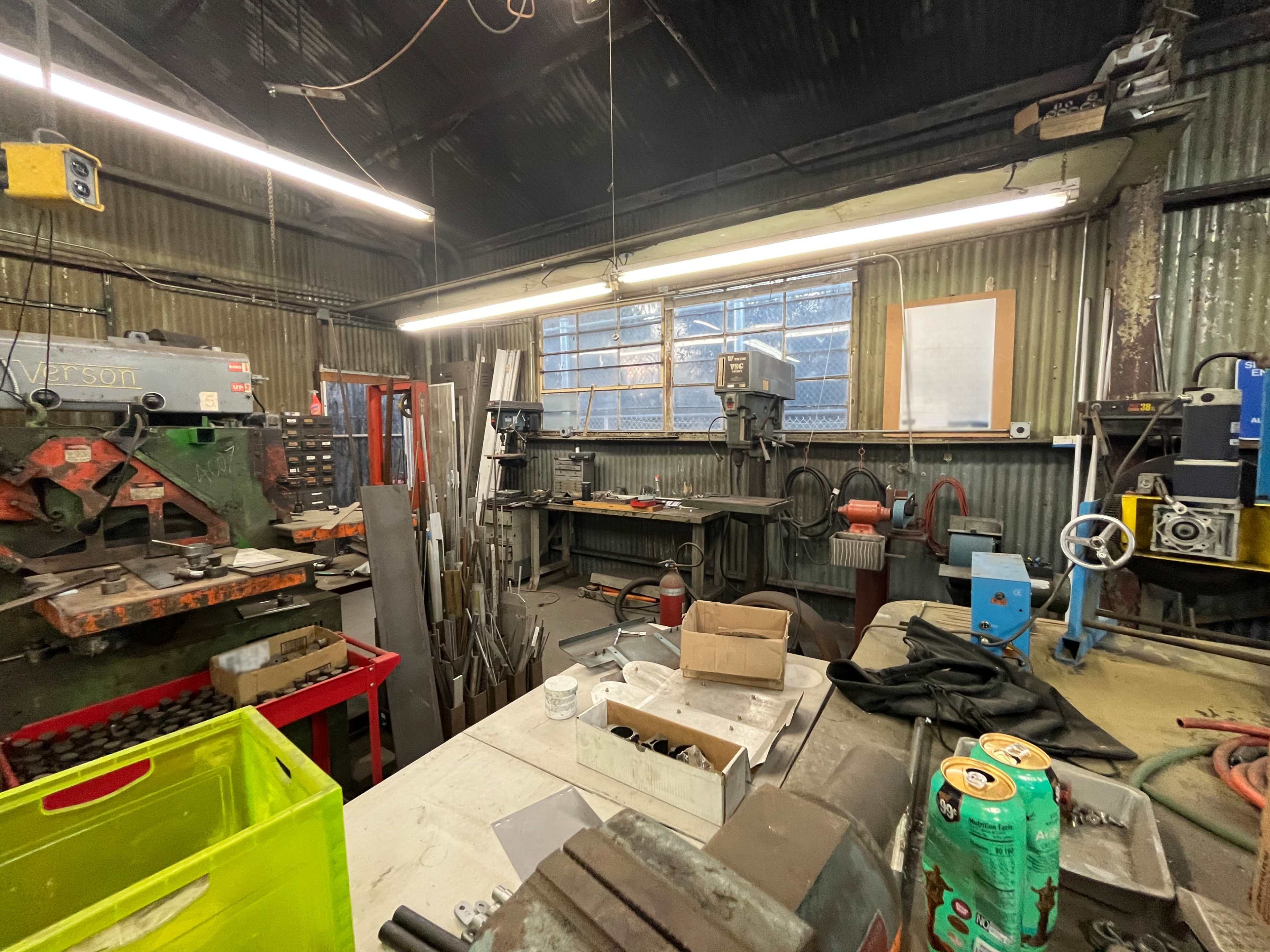 The image shows a cluttered workshop filled with various tools, machinery, and materials, illuminated by fluorescent lights.