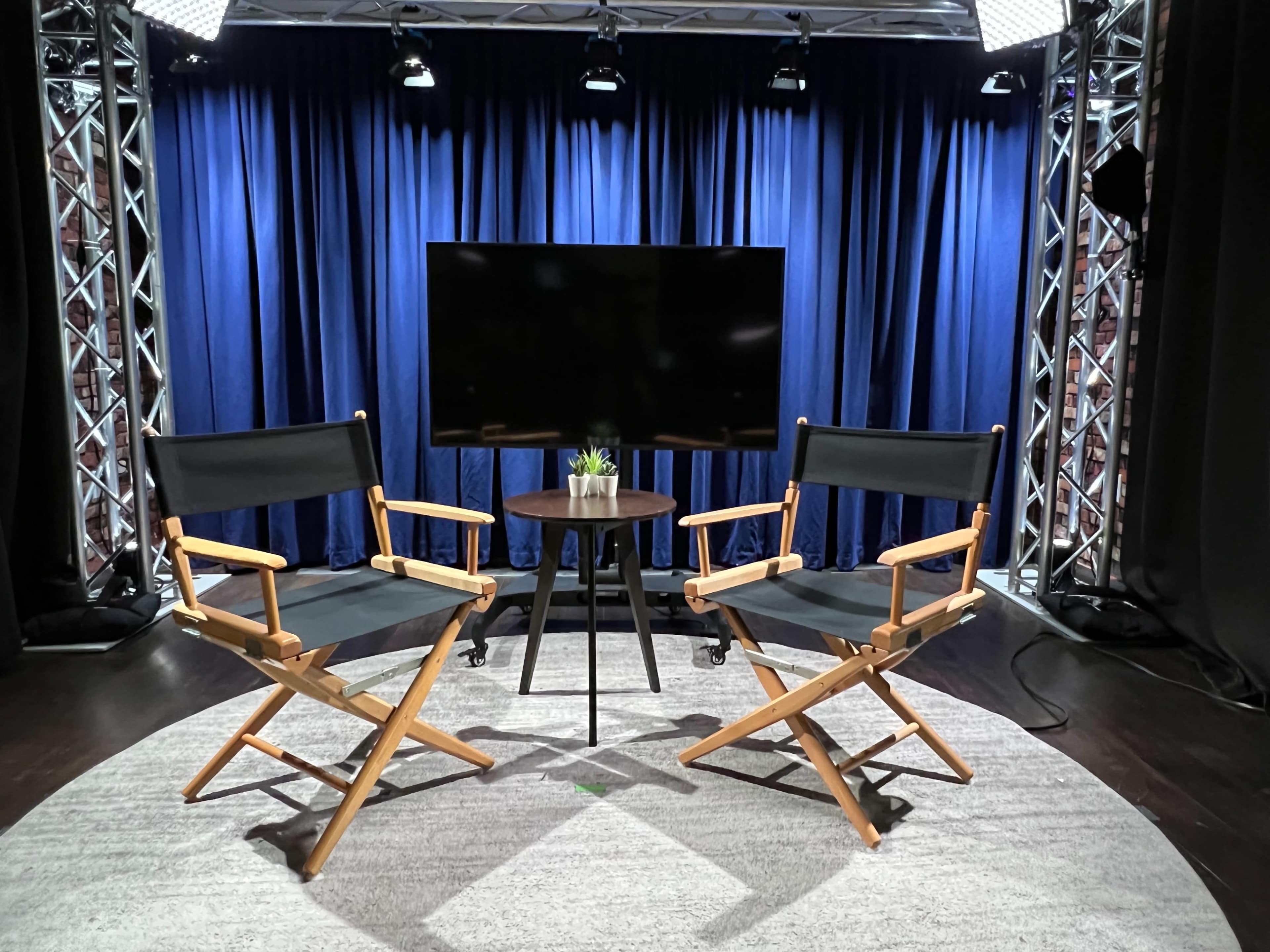 Two director's chairs placed in front of a large screen, with a small circular table and a potted plant between them, all set against a backdrop of blue curtains and studio lighting.