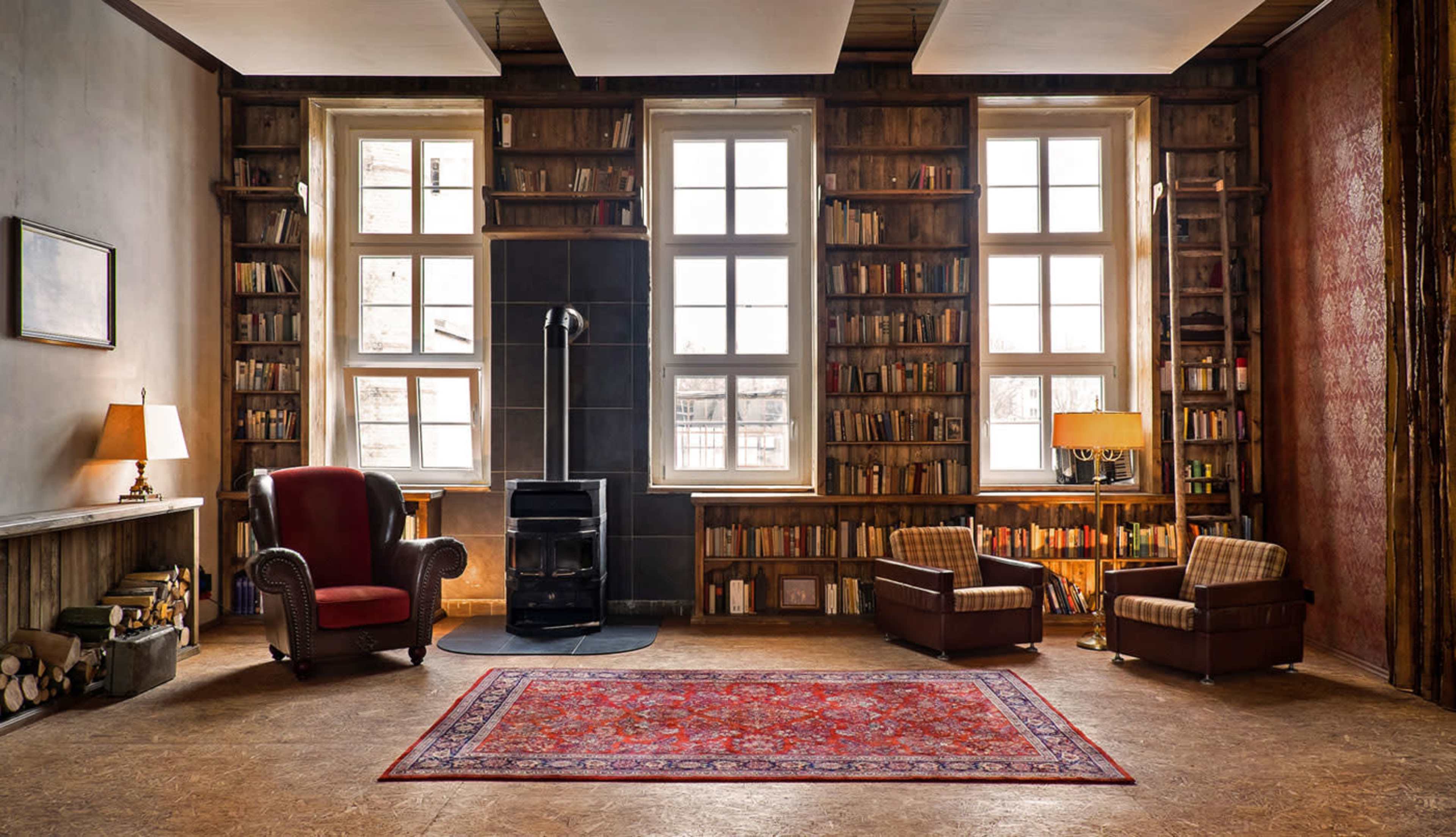 A cozy room features a red armchair and two plaid chairs, a small stove, and bookshelves filled with books along the walls.