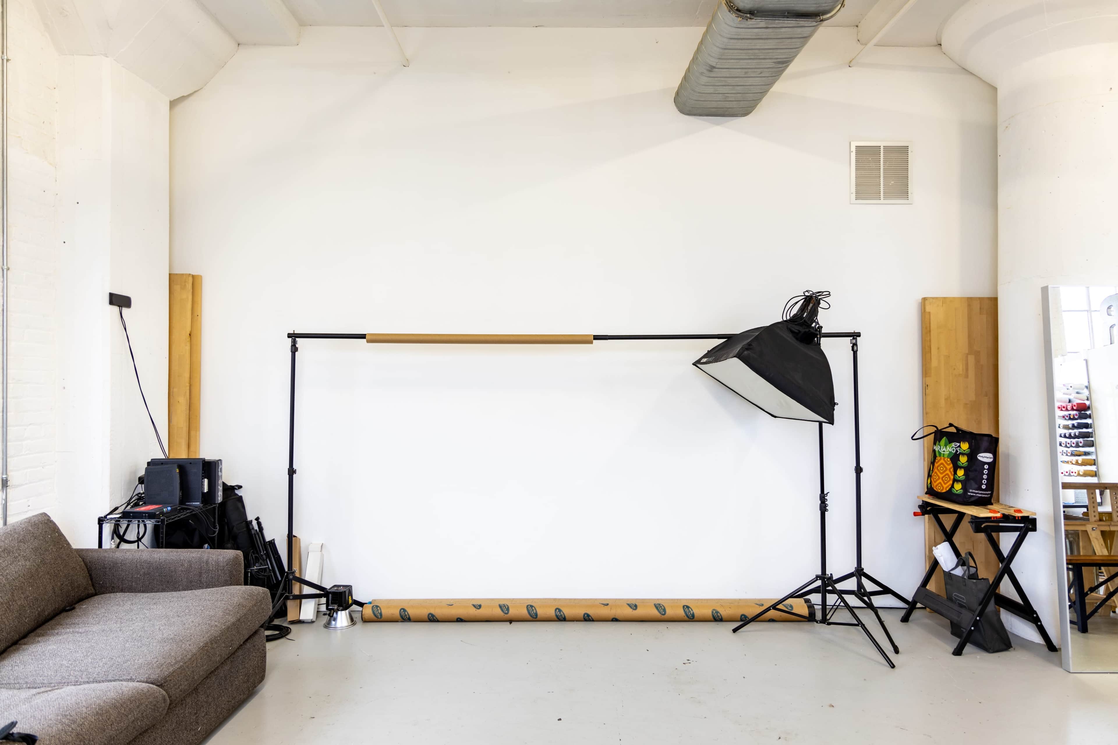 A minimalistic studio space features a gray couch, a lighting setup on a stand, and a vertical mirror against a white wall.