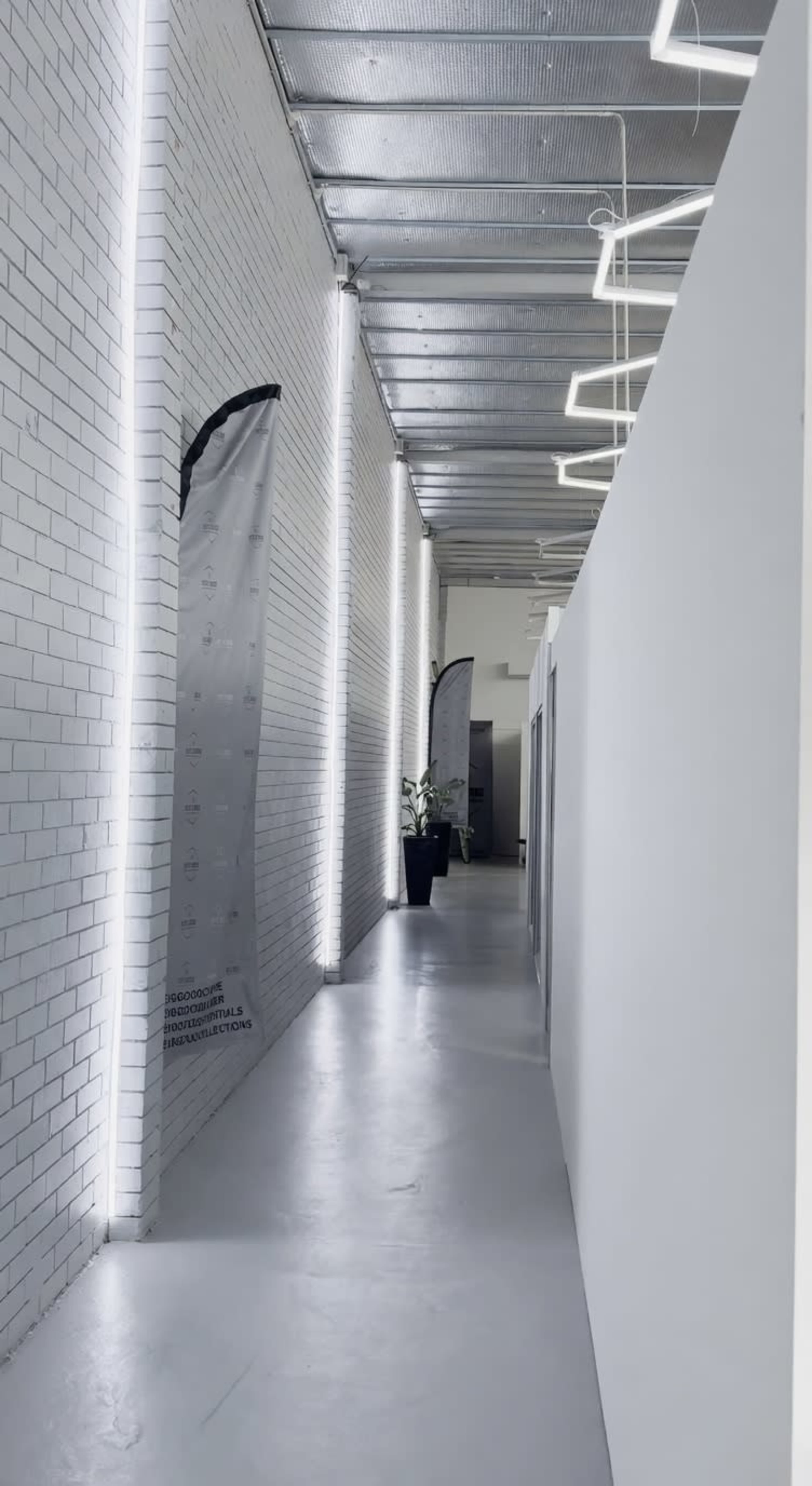 The image shows a long, narrow hallway with white brick walls, overhead lighting, and a few decorative elements including a plant in a pot.