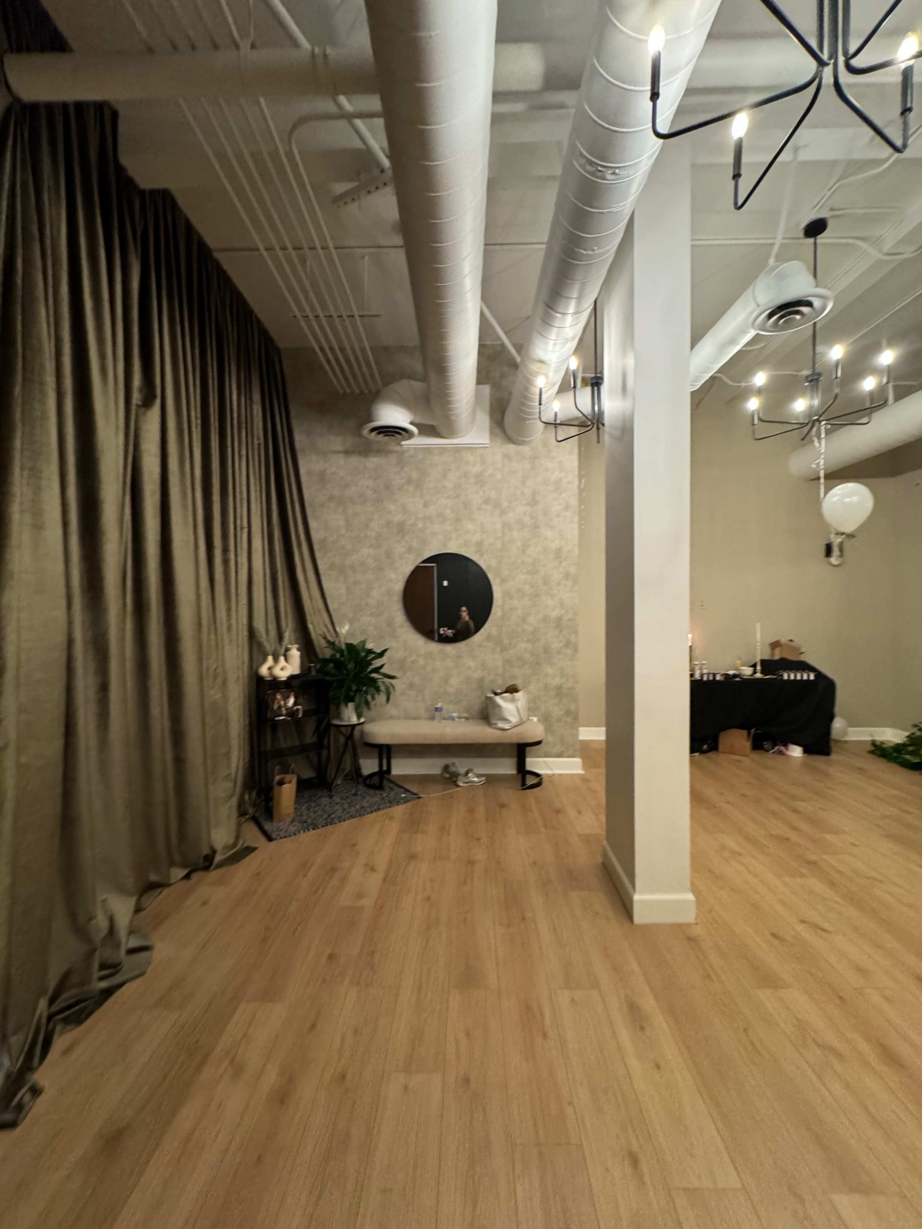 The image shows a spacious interior with wooden flooring, a circular mirror mounted on the textured wall, and a bench against a curtain, with party decorations in the background.