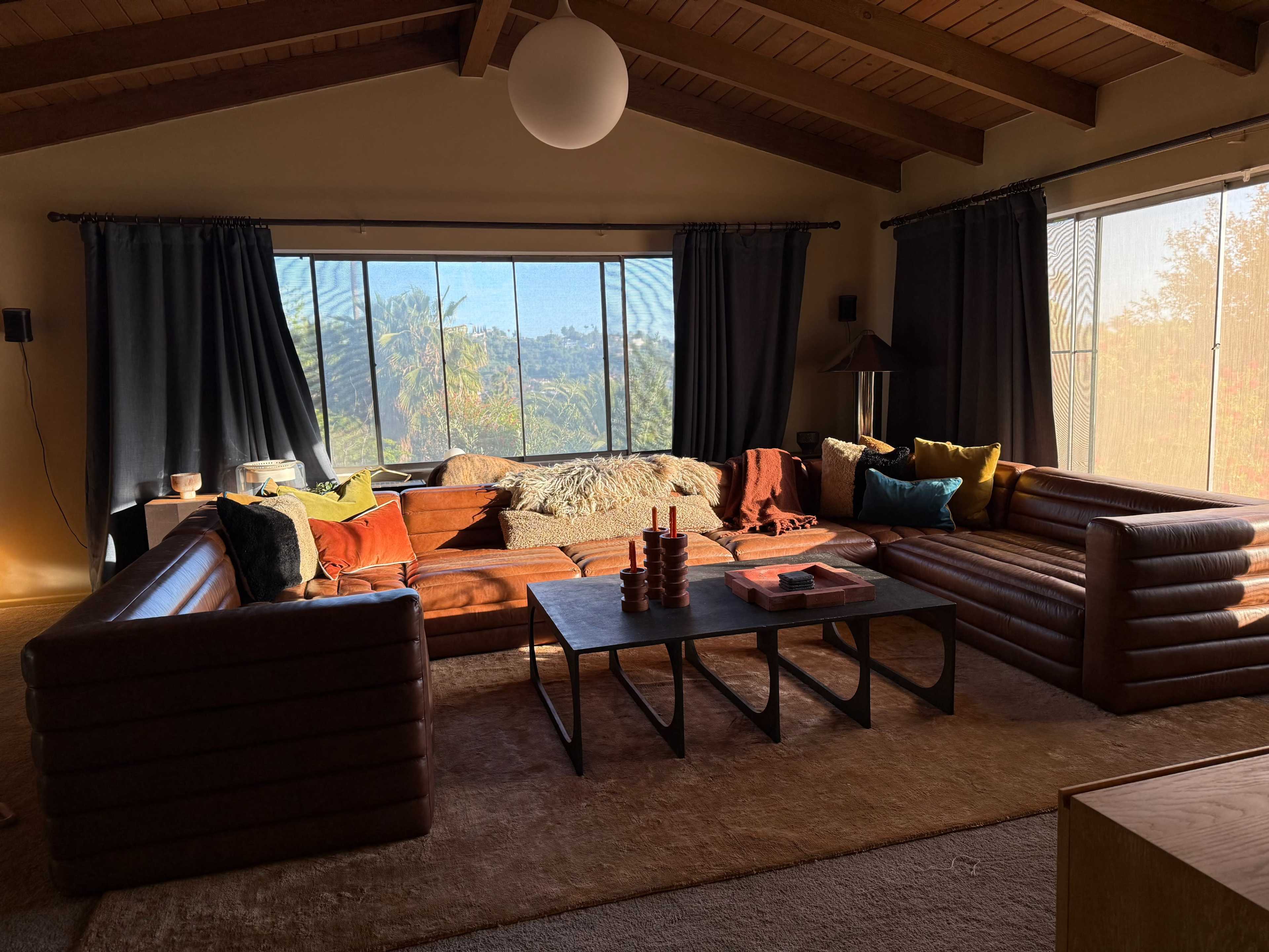 A spacious living room features a large brown sectional sofa, a coffee table, and large windows showcasing a view of palm trees and hills in the background.