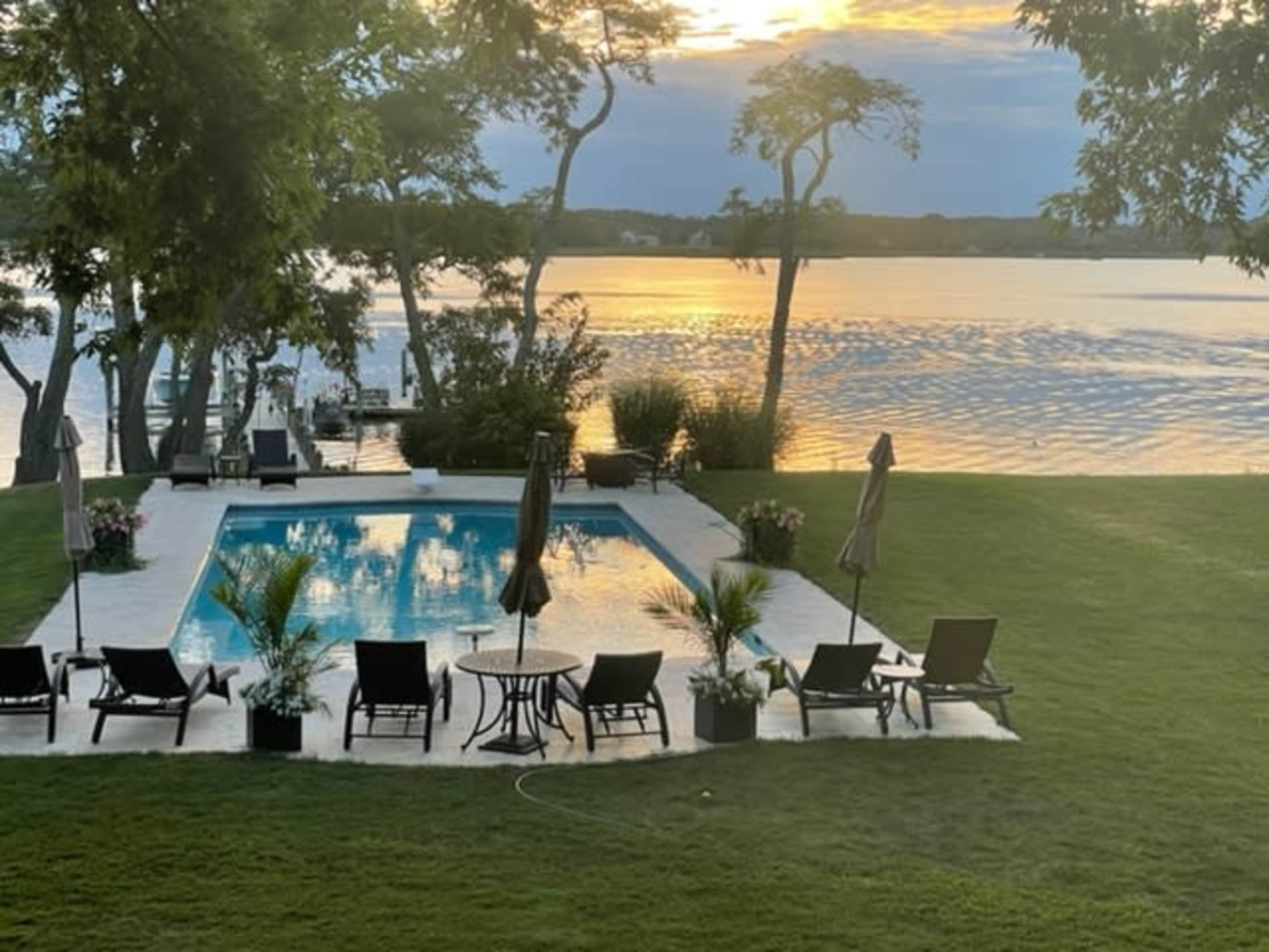 A swimming pool surrounded by lounge chairs and umbrellas, set against a lake with a sunset reflecting on the water.