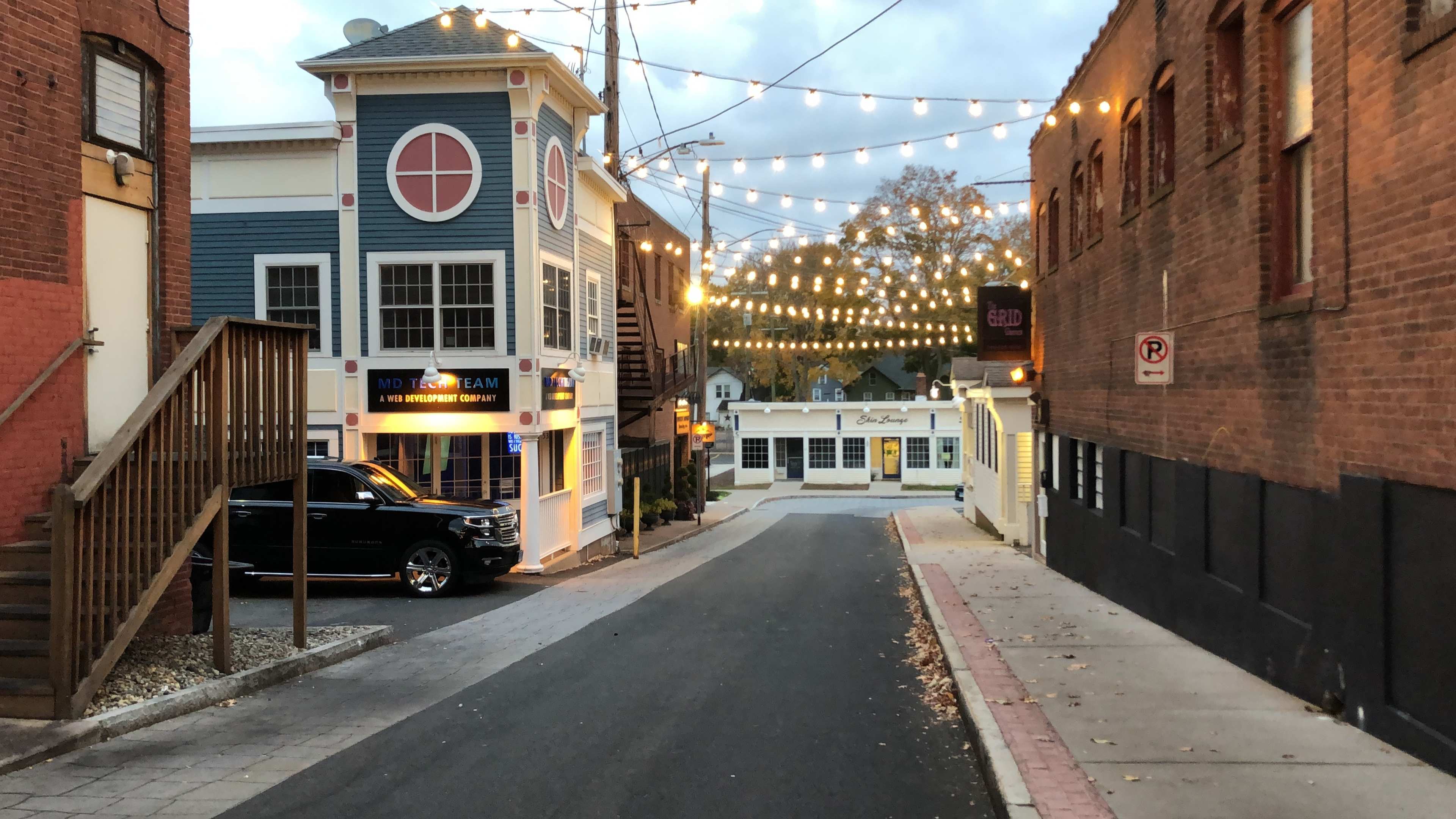 A narrow street lined with string lights features a blue building with round windows and a parked black SUV.