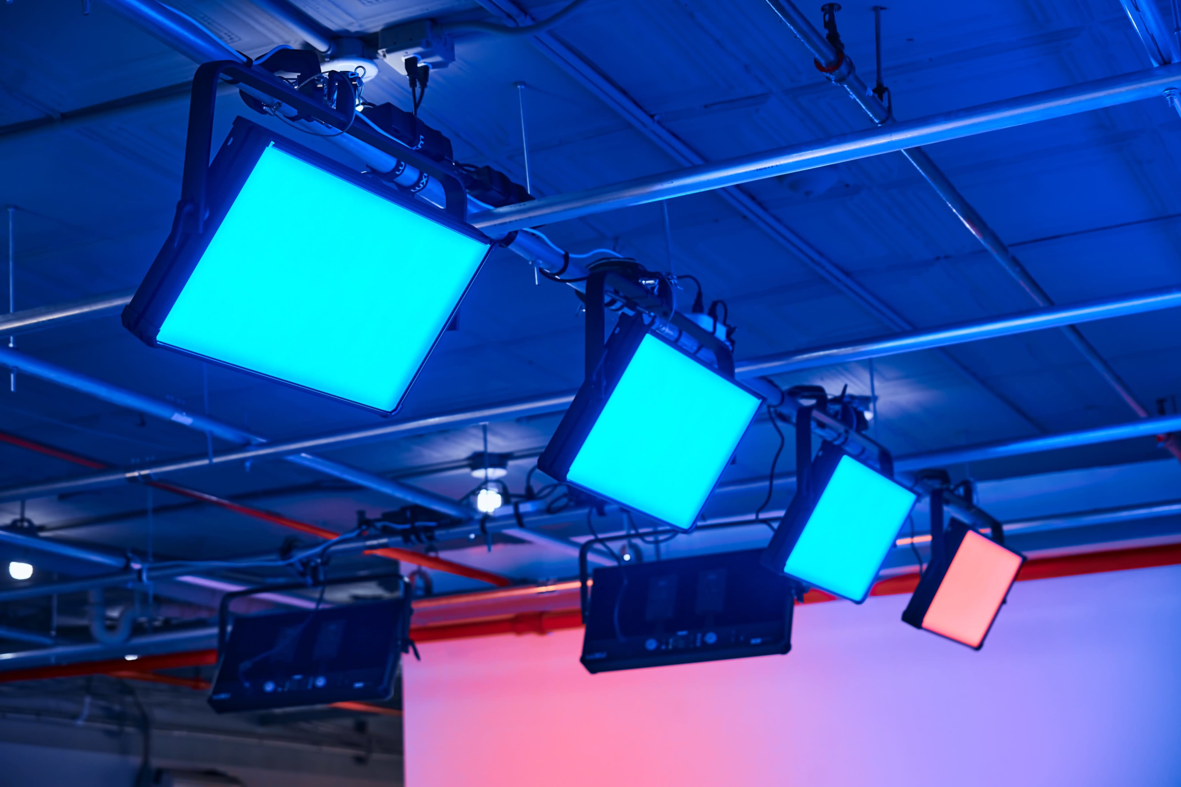Several rectangular LED lights in blue and red hues are suspended from a metal framework in a ceiling, illuminating a plain background.