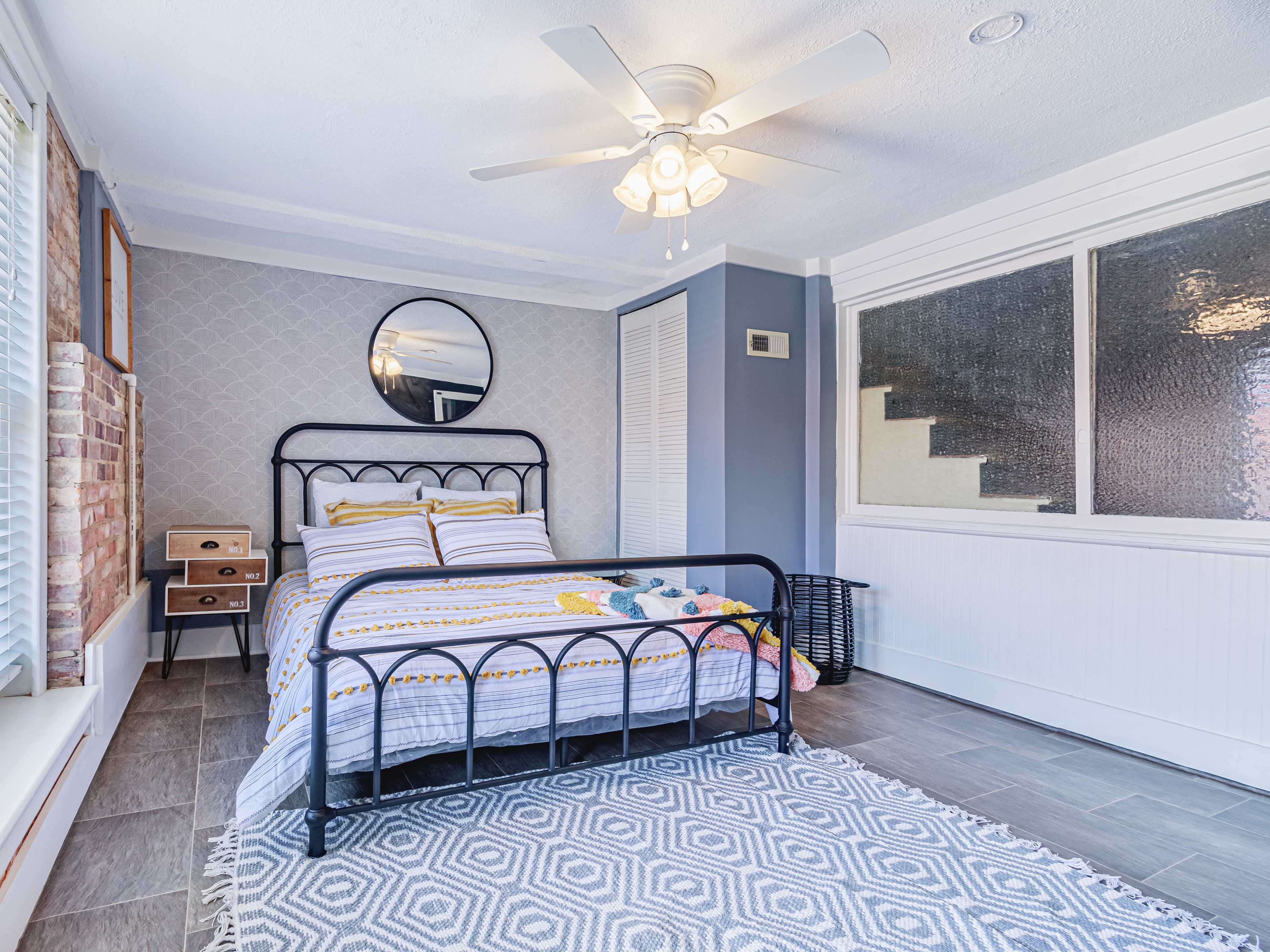 The image shows a bedroom featuring a black metal bed frame, a round mirror on the wall, and large windows with patterned curtains.