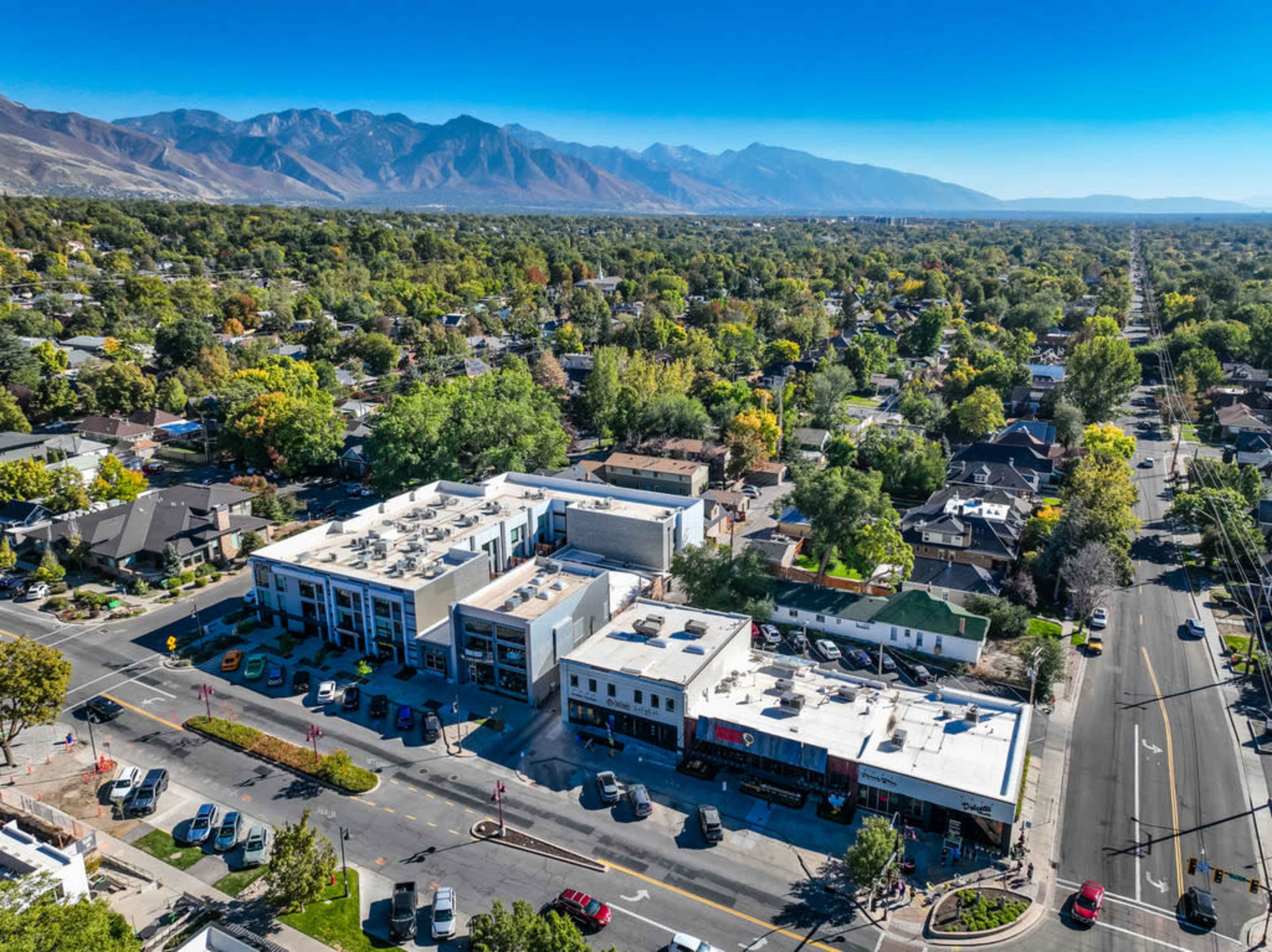 An aerial view of a suburban area with a multi-story building and several parked cars along a street, surrounded by greenery and mountains in the background.