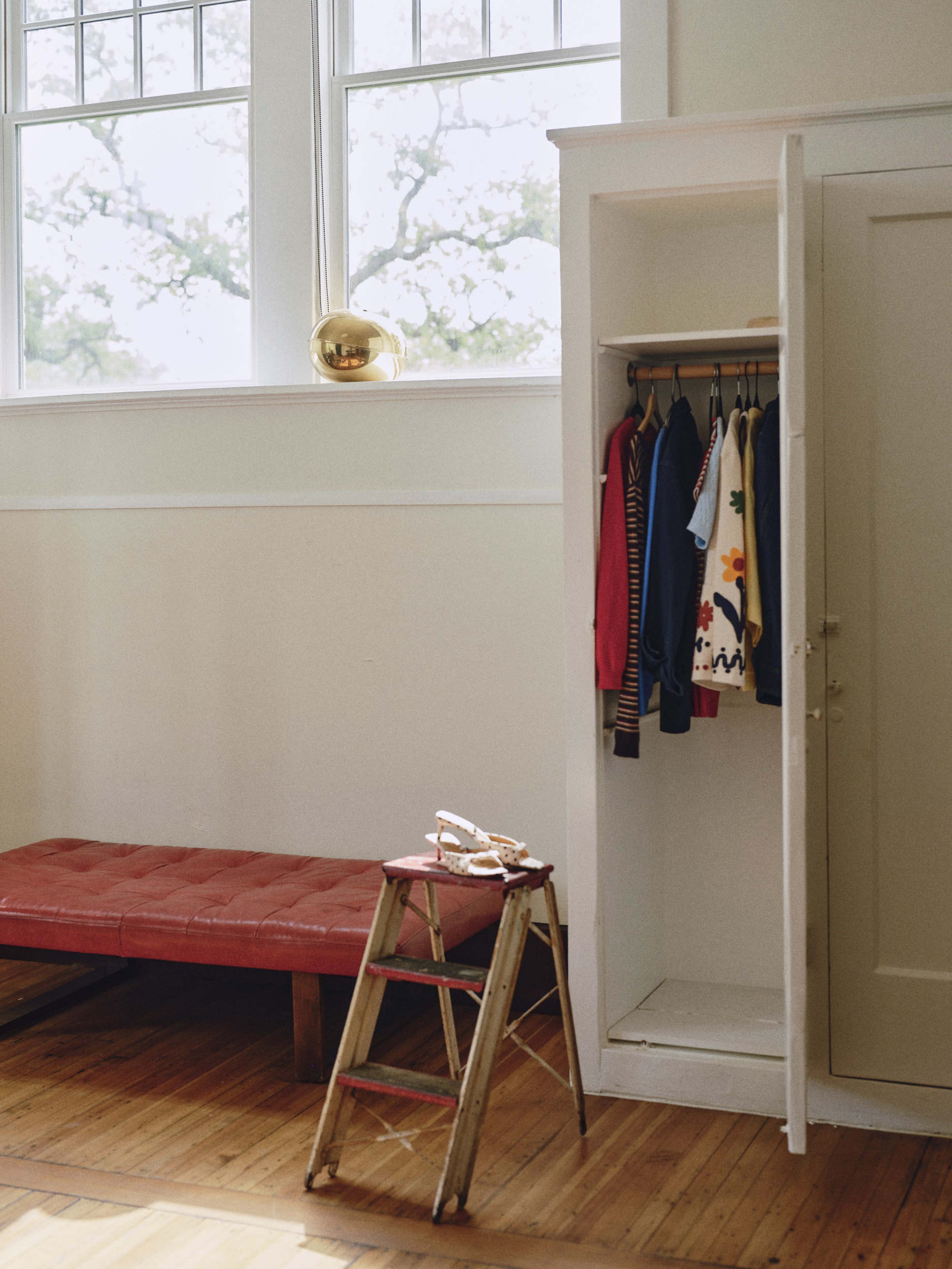 A white wardrobe with hanging clothes is positioned next to a red bench and a wooden step ladder in a bright room with large windows.