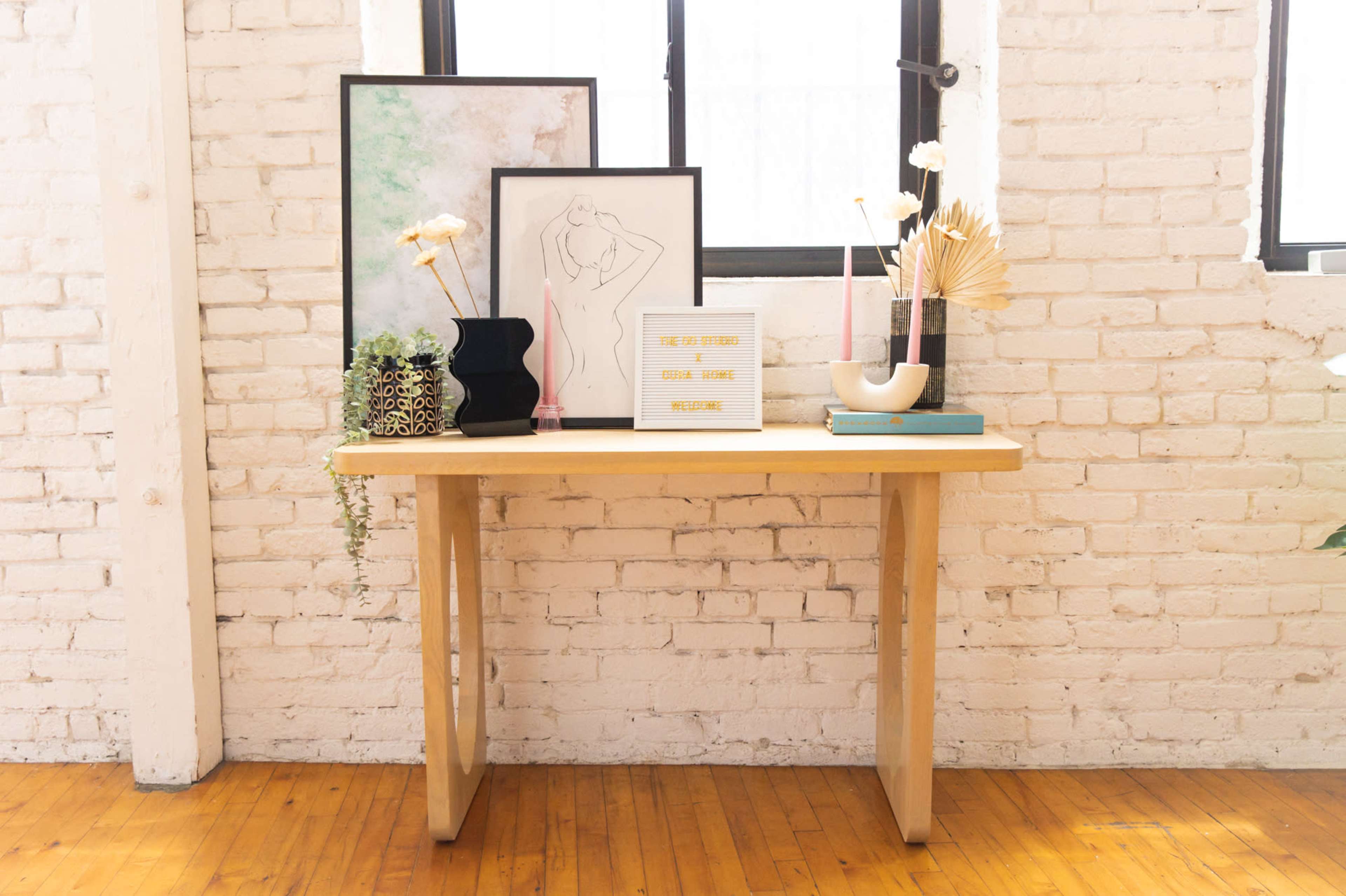 A wooden table displays various framed artworks, decorative vases, and a plant in a bright, minimalist room.