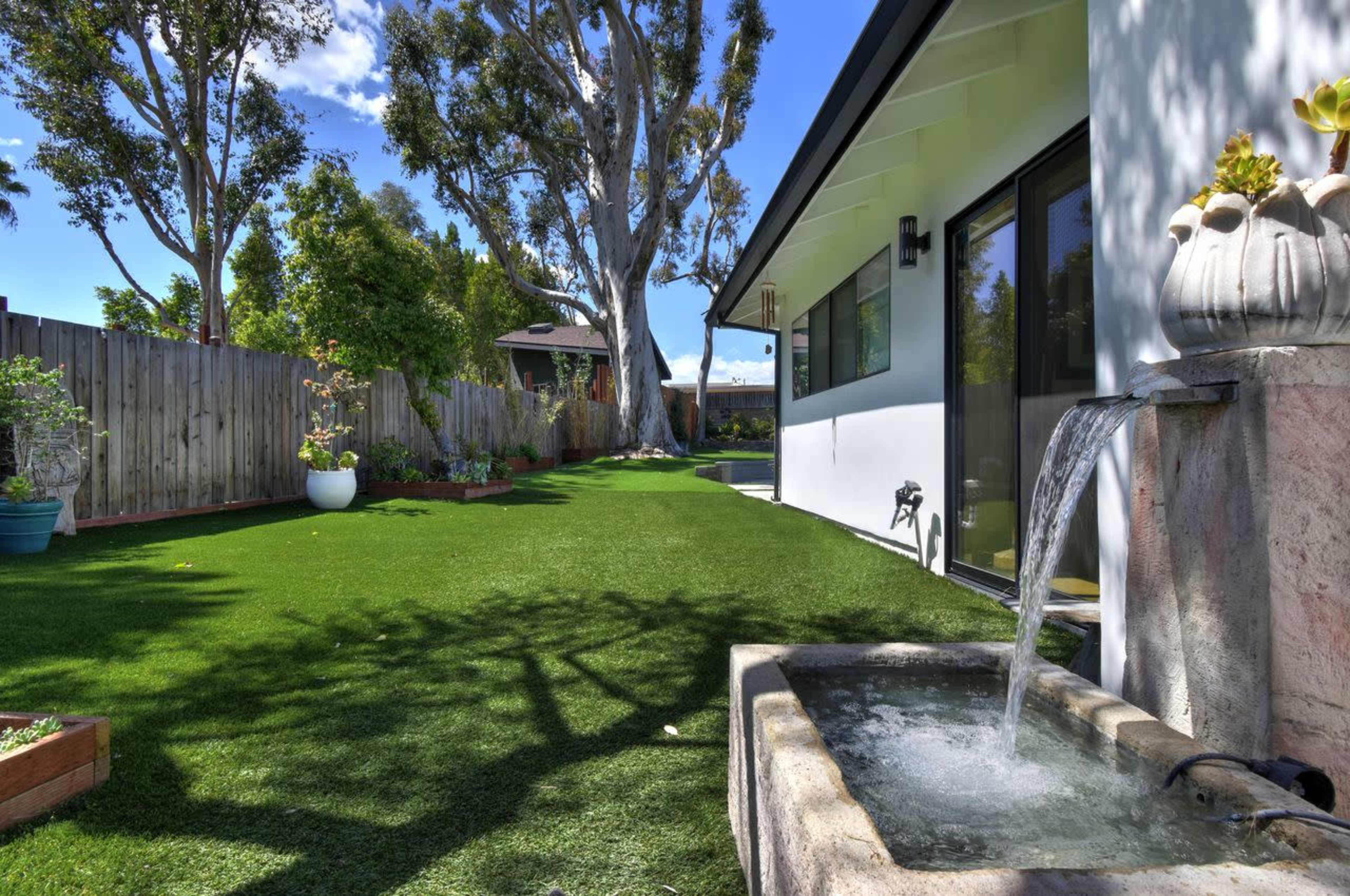 A landscaped yard features synthetic grass, a stone water fountain, and well-maintained plant beds alongside a house.