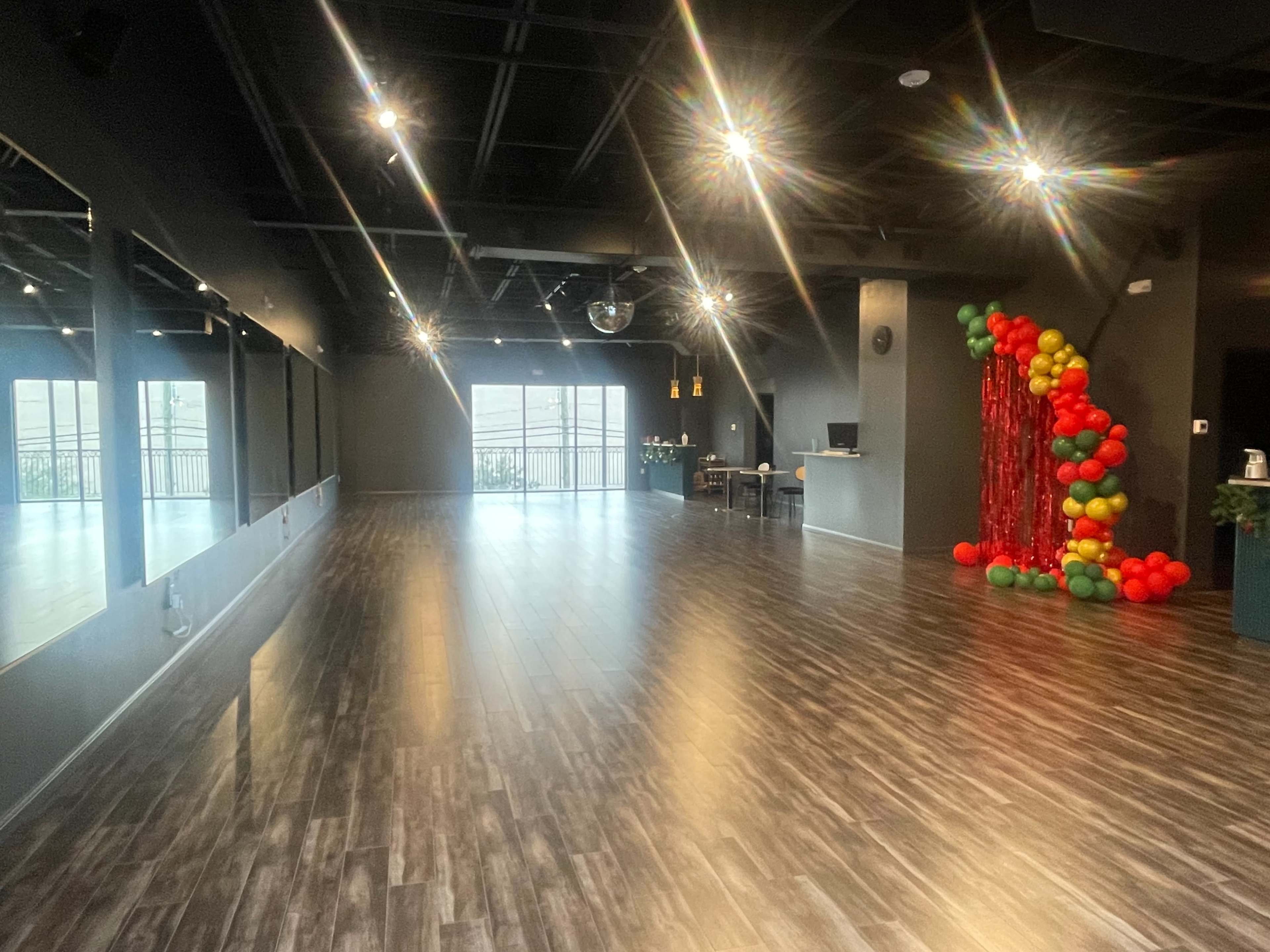 A spacious, well-lit studio features mirrored walls, polished wooden floors, and a decorative arrangement of colorful balloons on one side.