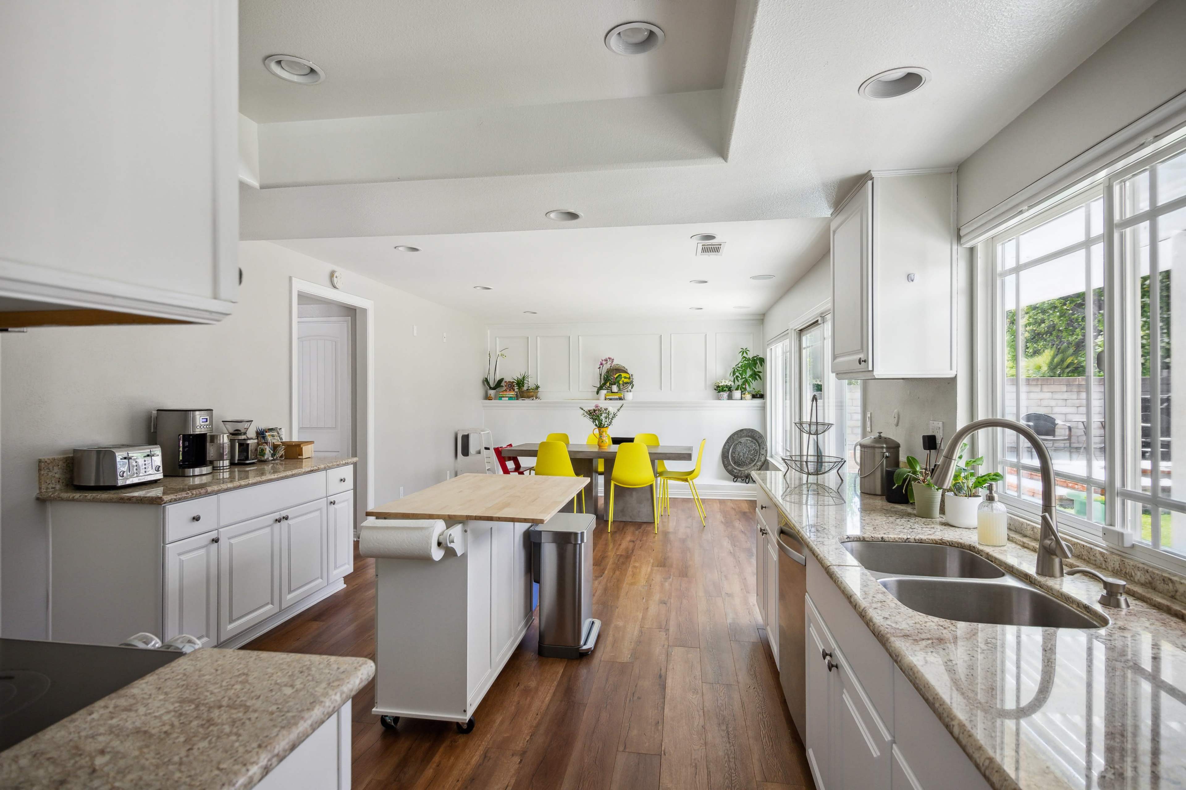 A modern kitchen features white cabinets, a central island, and a dining area with yellow chairs near large windows overlooking an outdoor space.