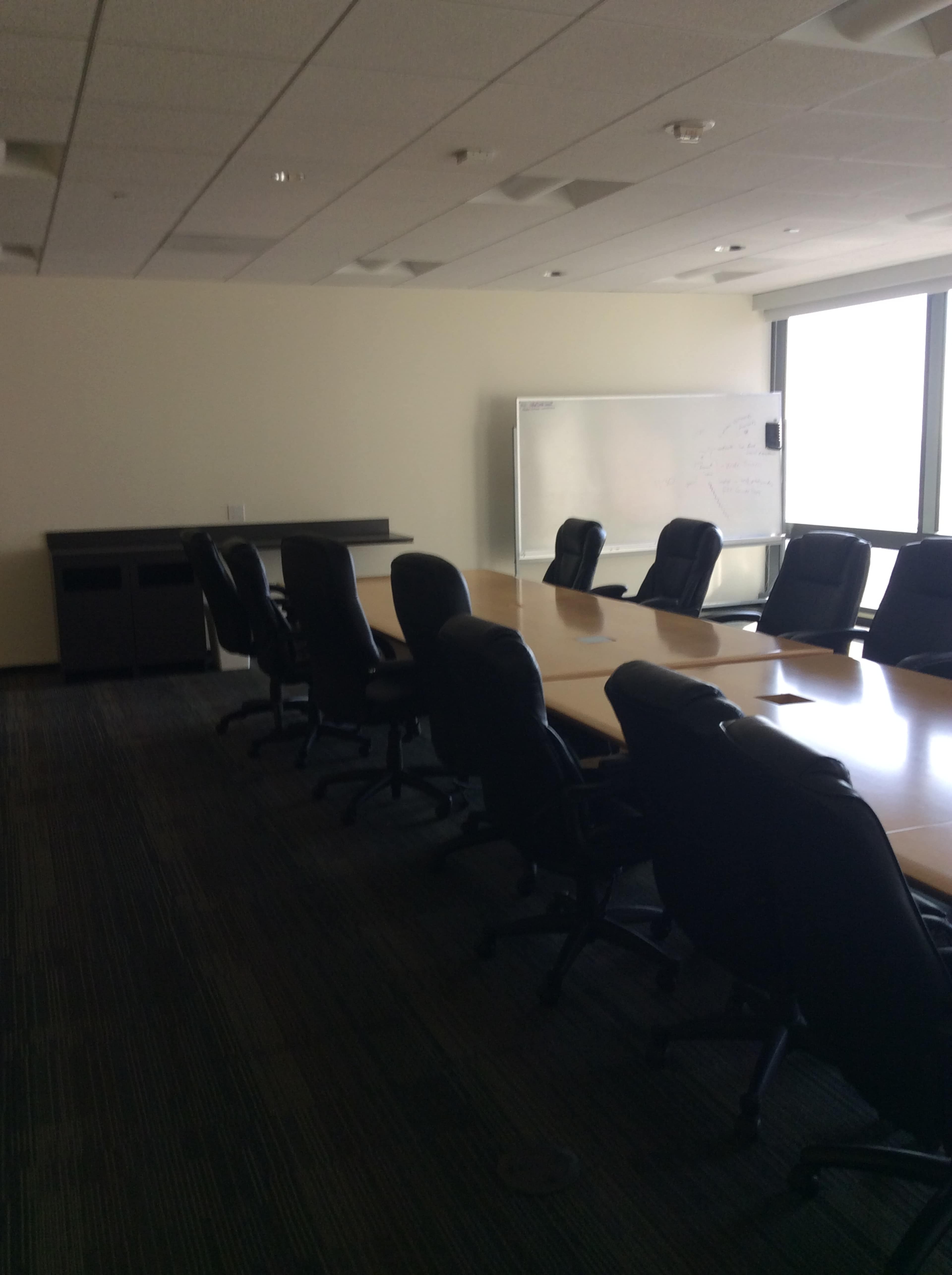 A conference room features a long table surrounded by black chairs, with a whiteboard positioned against a wall and large windows allowing natural light.