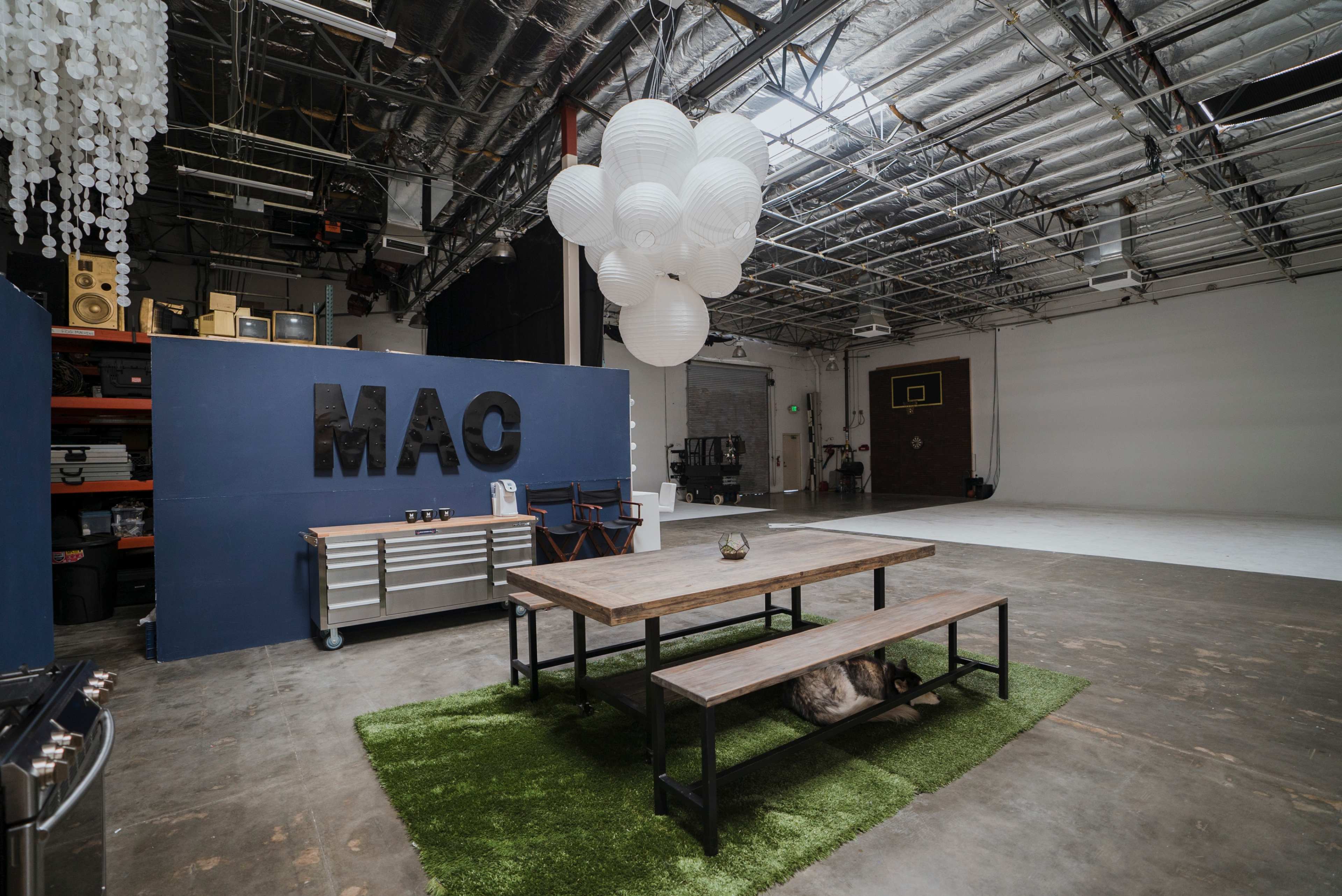 A spacious indoor studio with a table and benches on a green rug, a blue wall with the letters "MAC," and hanging white lanterns above.