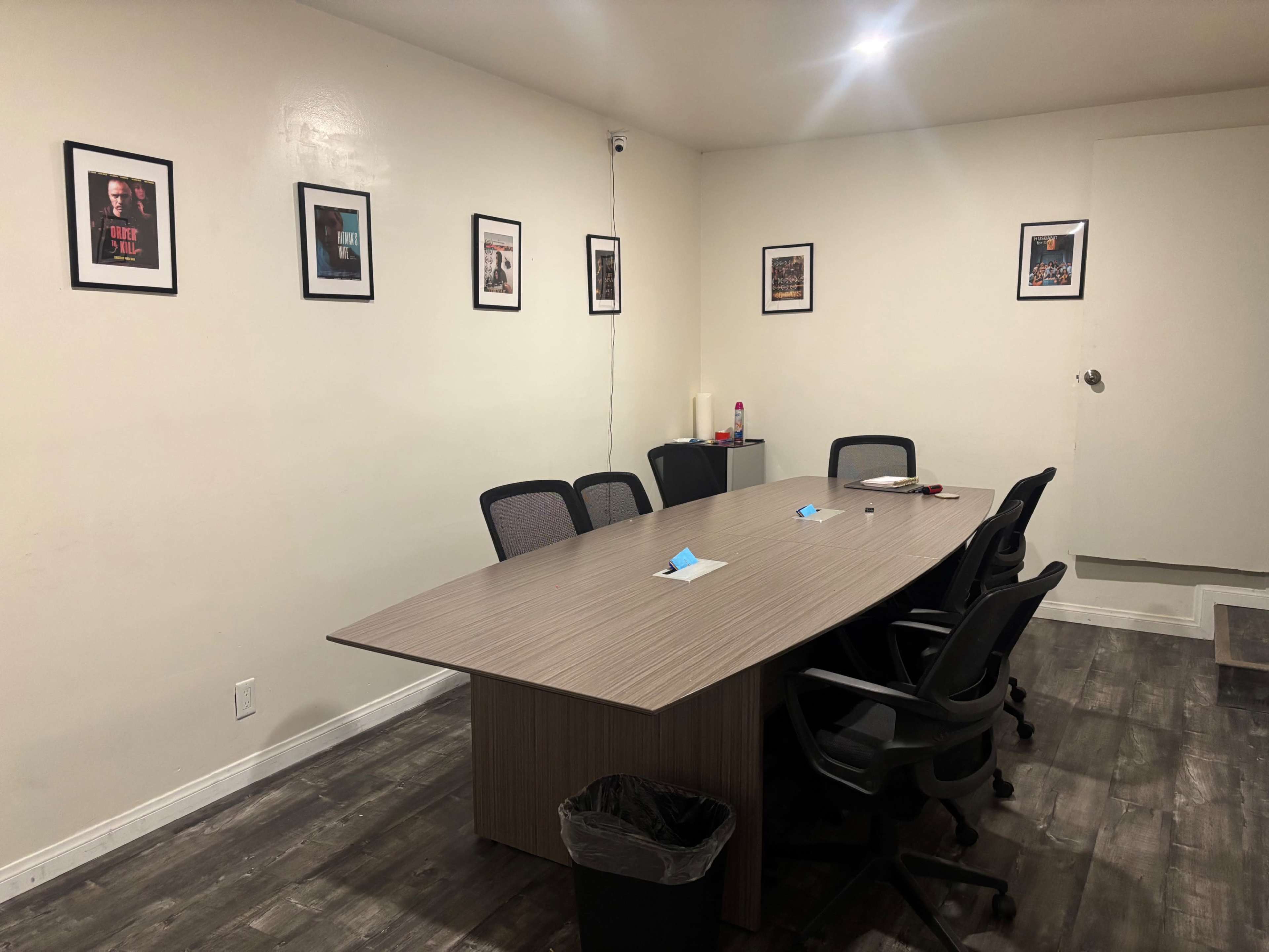 The image shows a conference room with a large table surrounded by chairs, featuring framed posters on the walls and a closed door in one corner.