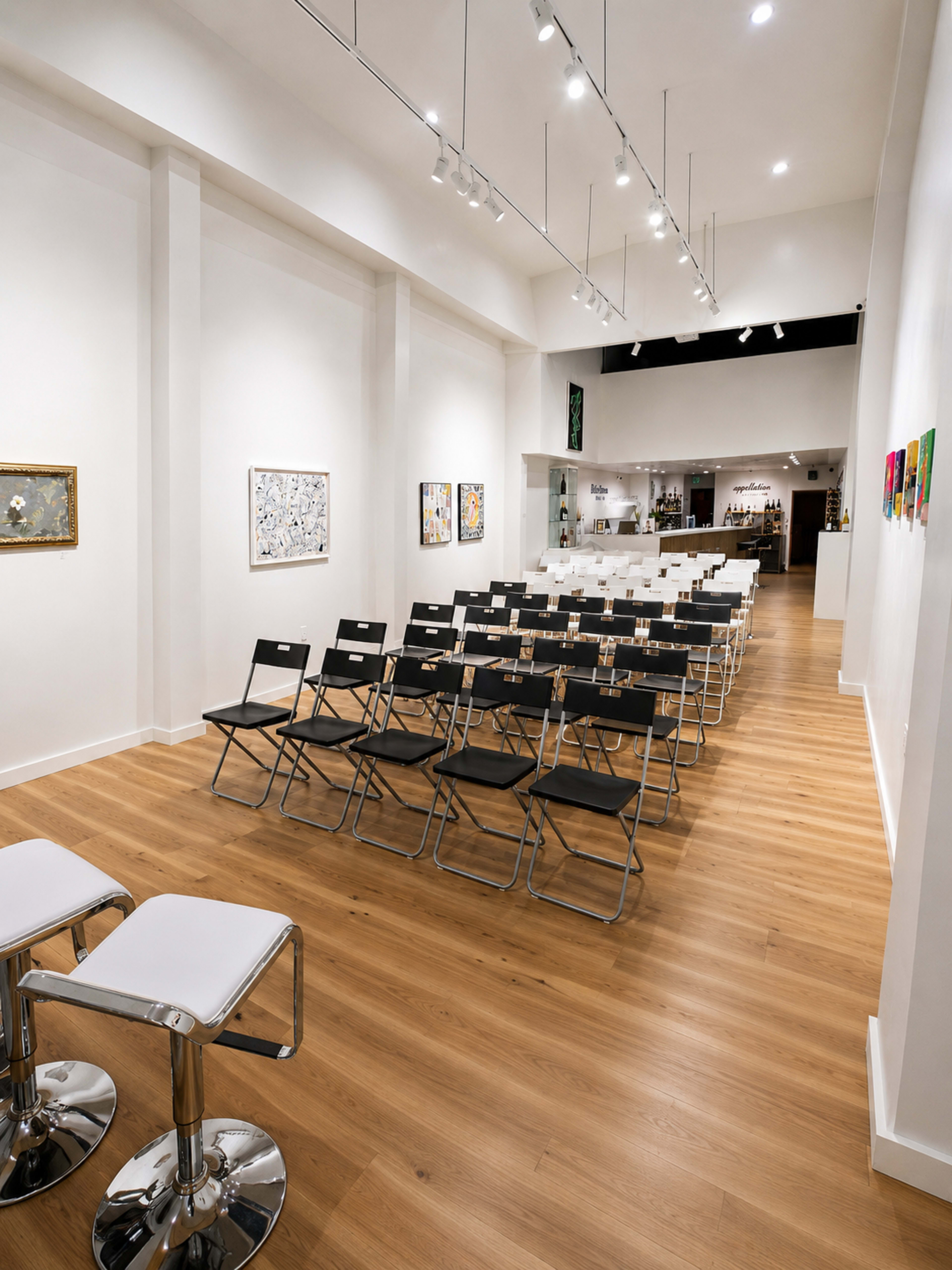 The image shows a spacious art gallery or event space featuring rows of black and white folding chairs, with artwork displayed on the white walls.