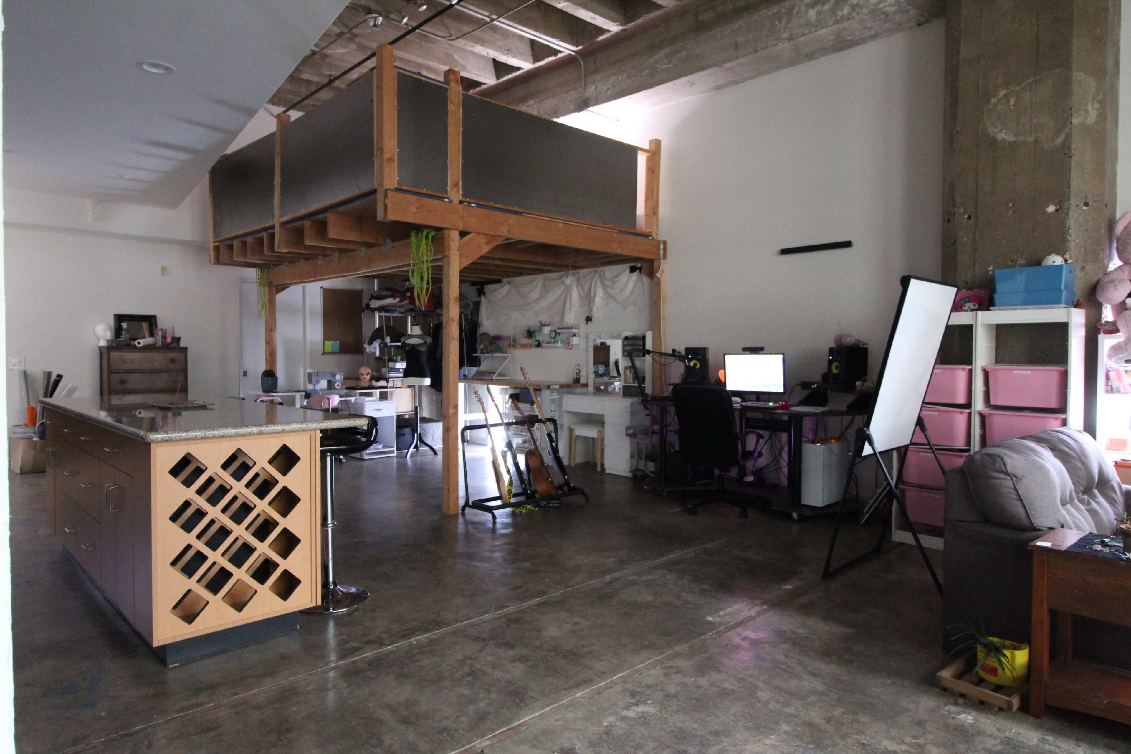 The image shows an open-concept loft space featuring a raised sleeping area, a kitchen island with a decorative panel, an office setup, and casual seating.