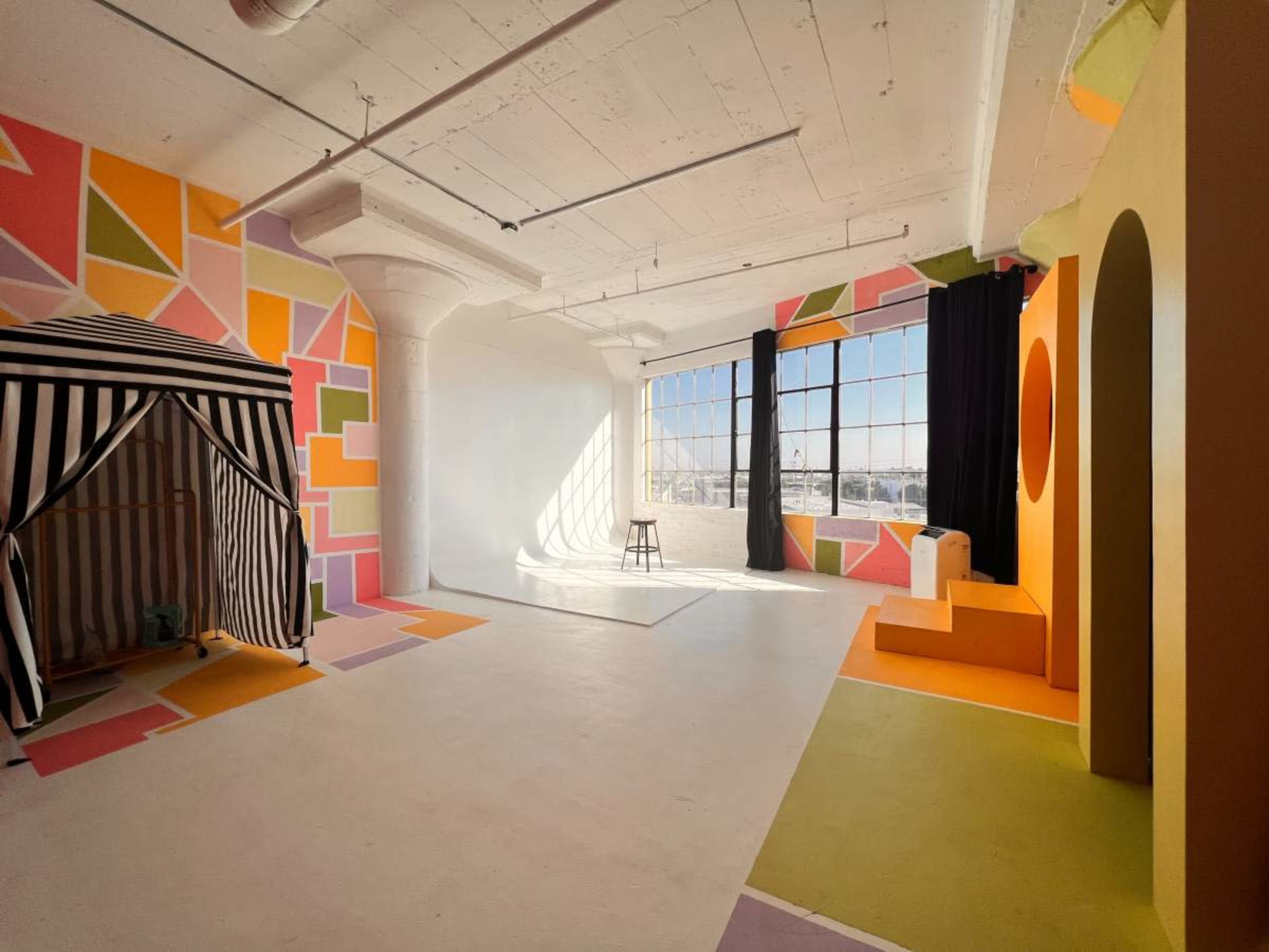 A brightly colored studio space featuring geometric wall patterns, a black and white striped tent, a stool, and various shaped structures in orange and yellow.
