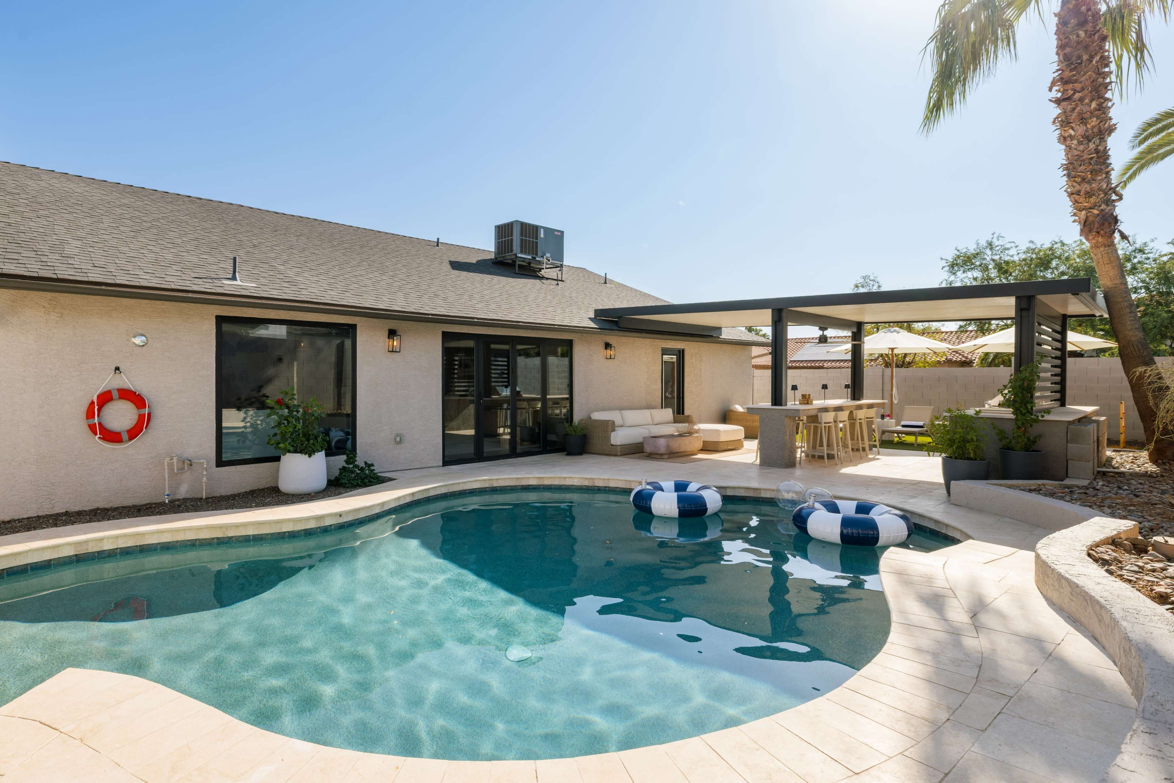 A backyard features a swimming pool with two blue and white floats, surrounded by a patio and palm trees.