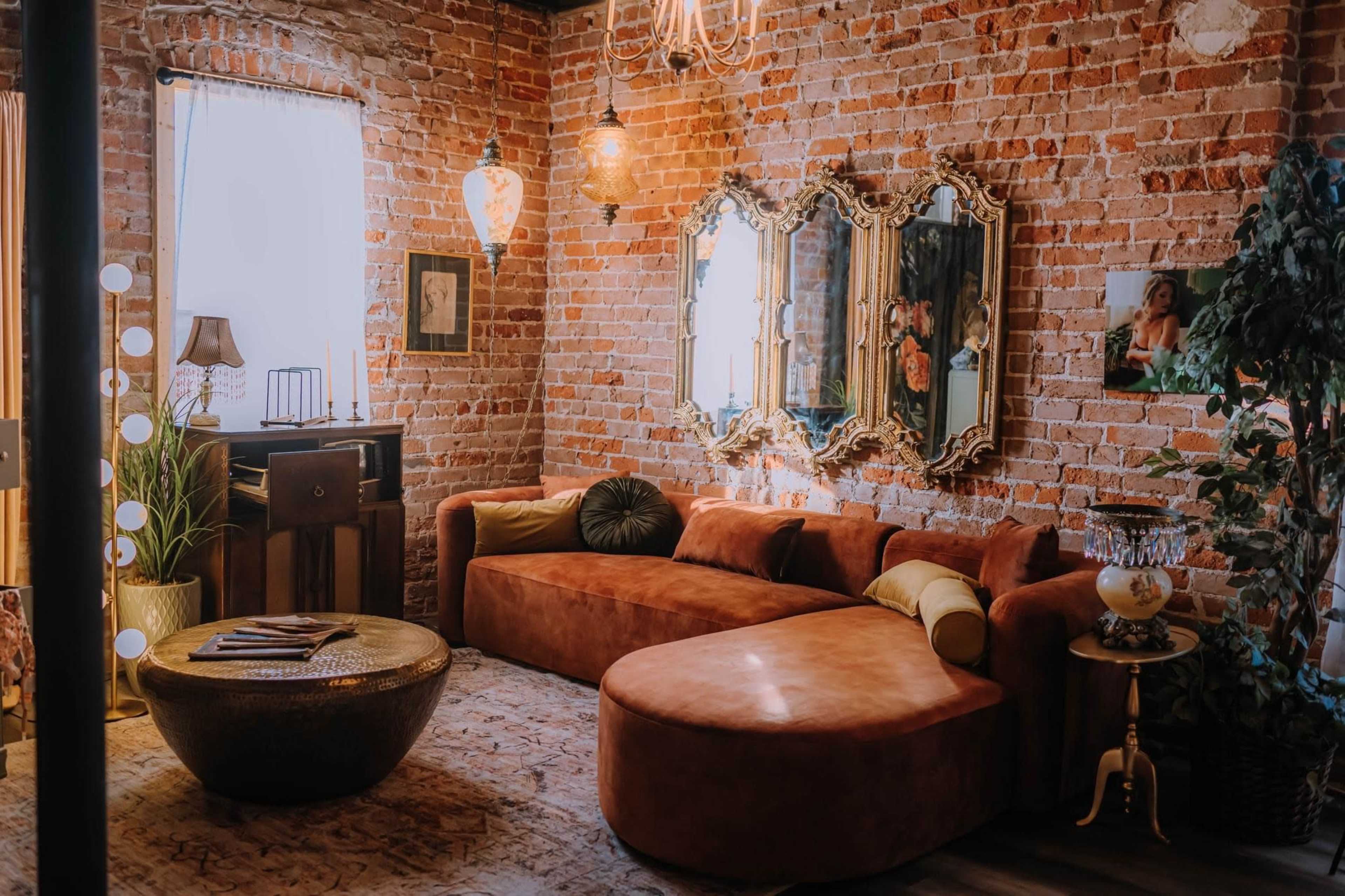 A cozy living room with exposed brick walls, a curved orange sofa, a round coffee table, and elegant mirrors reflecting warm lighting.