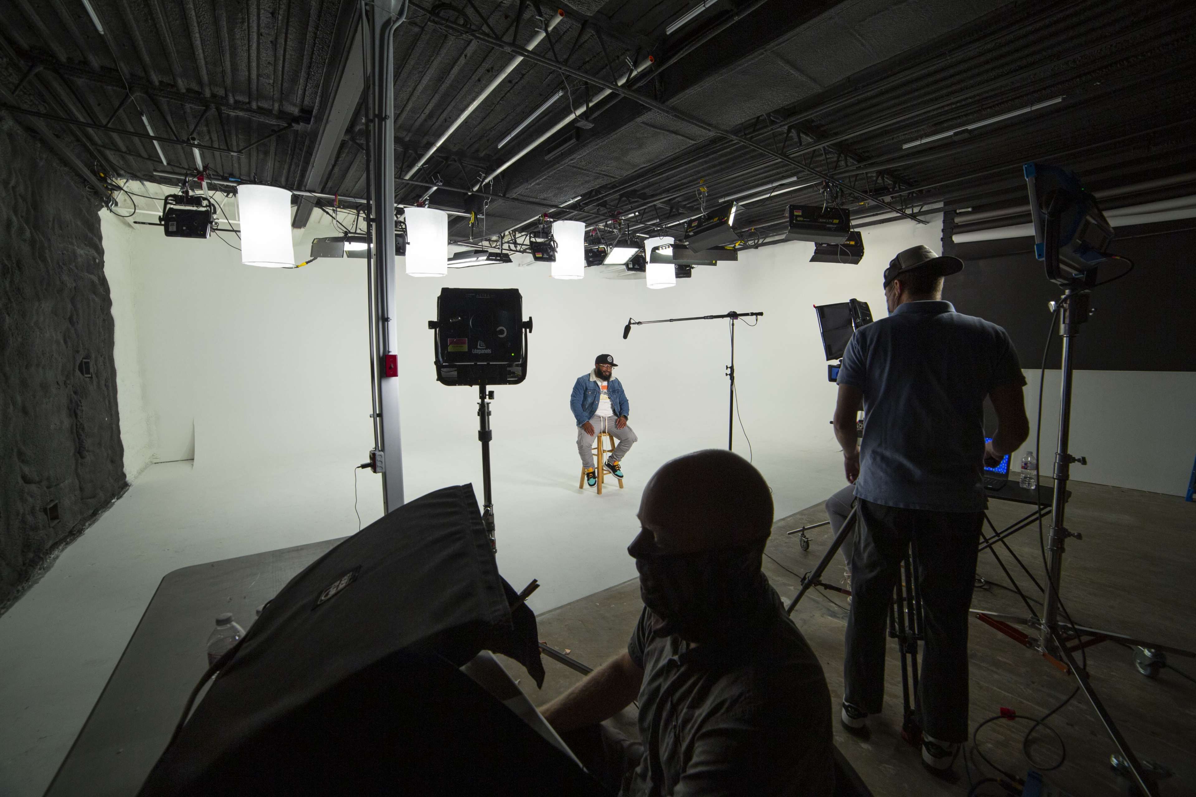 A film studio set is shown with a man sitting on a stool in front of a blank backdrop, while crew members operate cameras and lighting equipment.