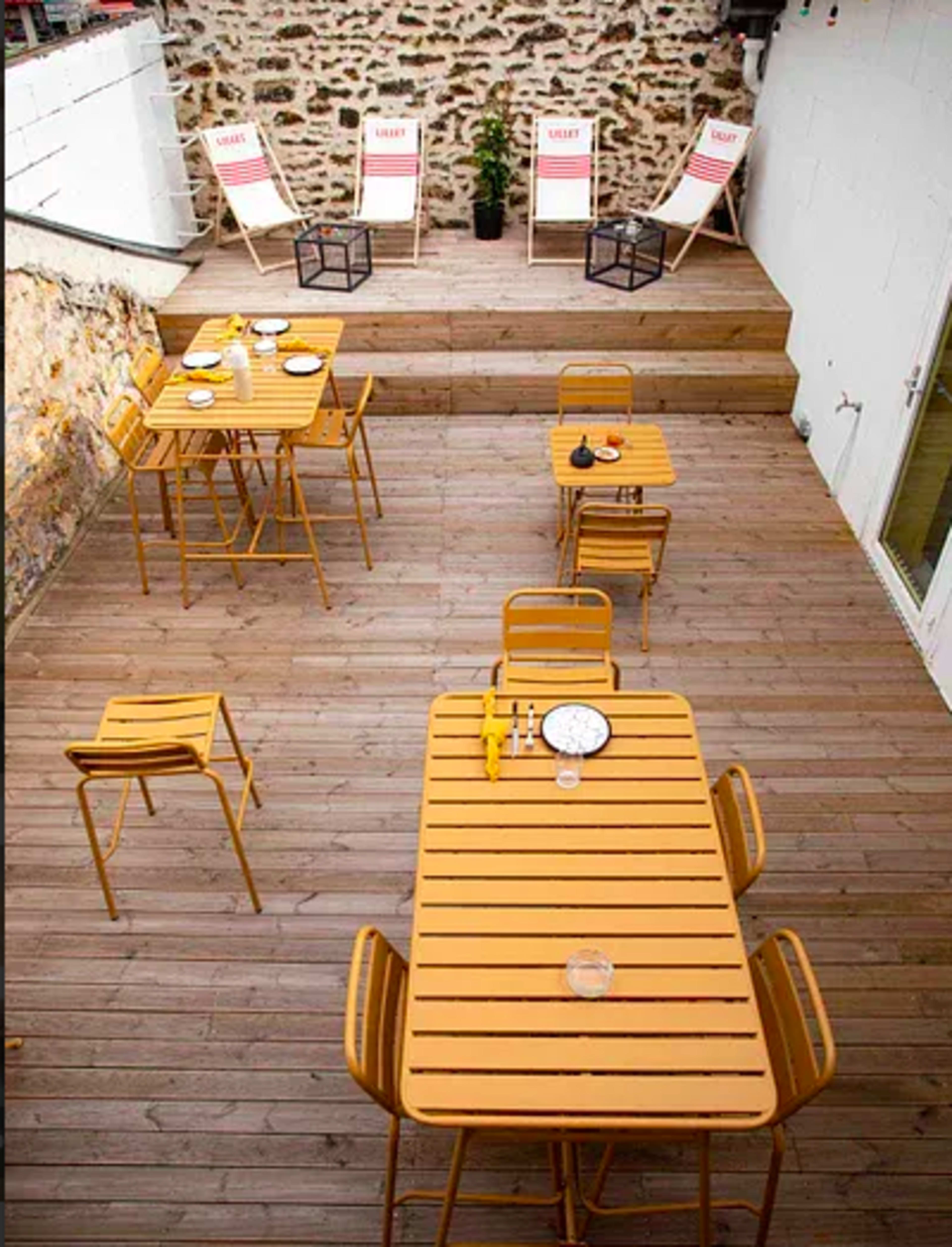 A wooden deck area with several yellow metal tables and chairs, accompanied by deck chairs in the background against a stone wall.