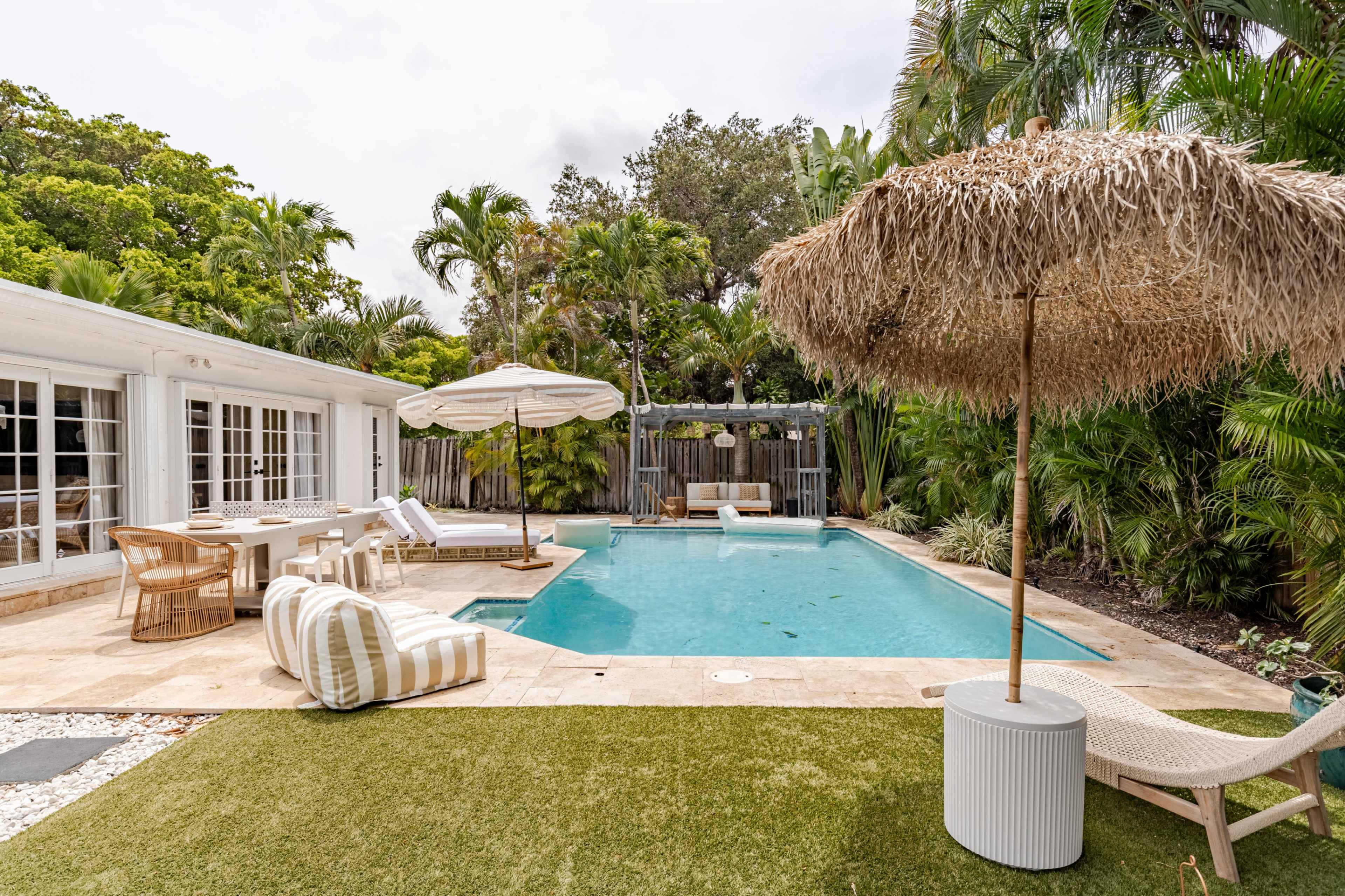 The image shows a landscaped backyard with a swimming pool, surrounded by palm trees and outdoor furniture, including a straw umbrella and lounge chairs.