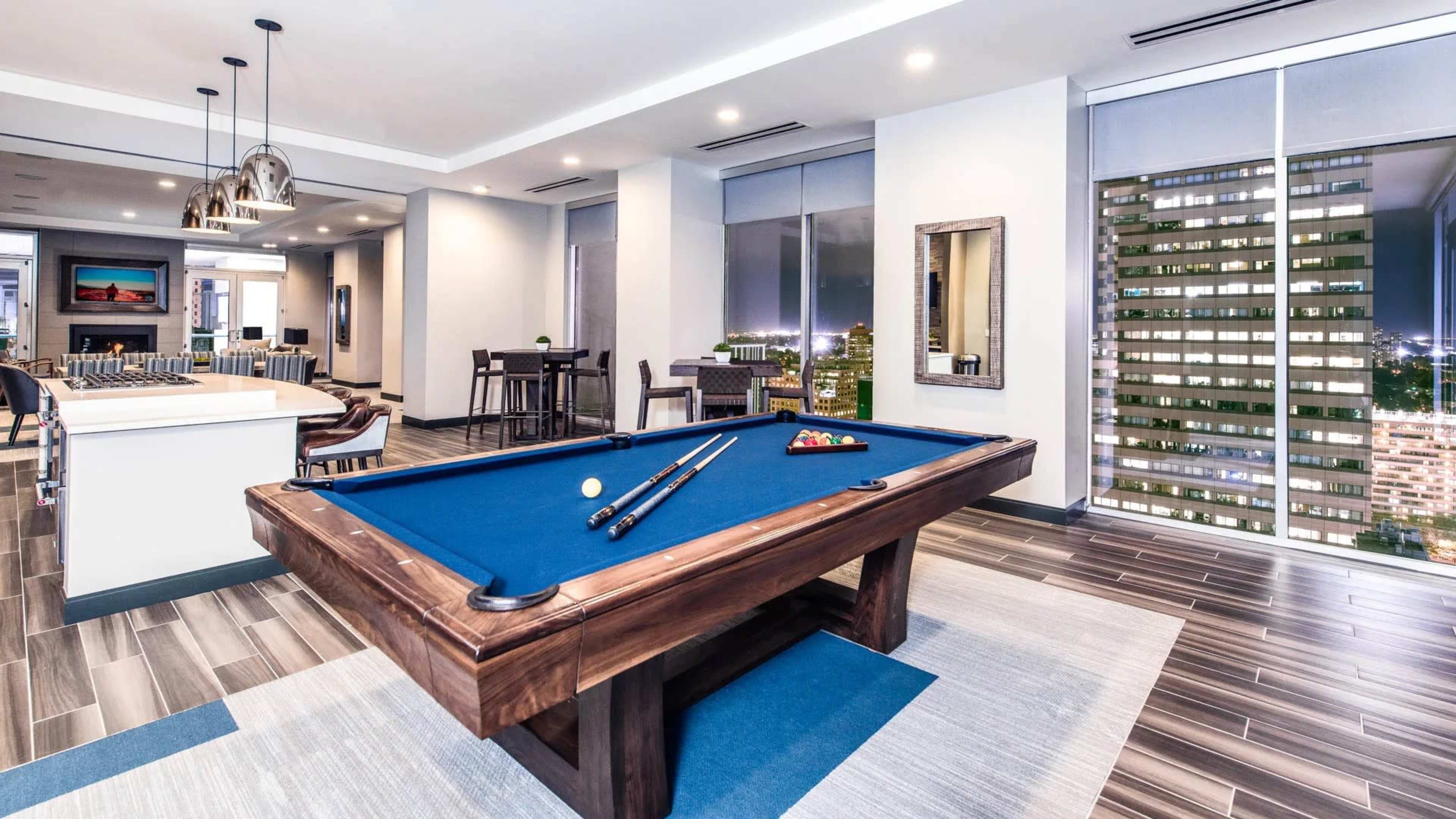 A well-lit game room features a blue pool table with cues and balls prepared for play, surrounded by modern furniture and large windows showing a city skyline.
