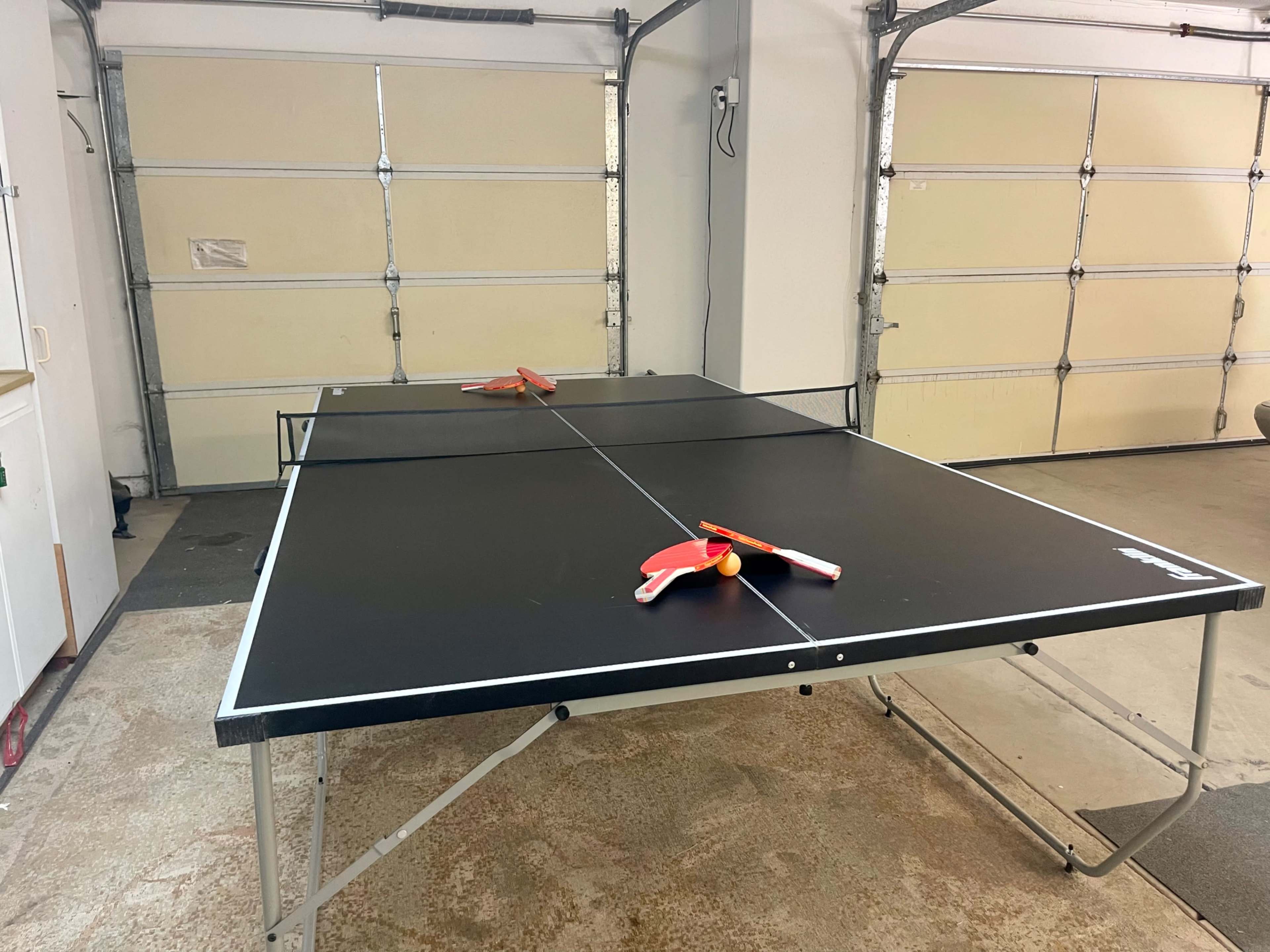 A black ping pong table with paddles and a ball is set up in a garage.