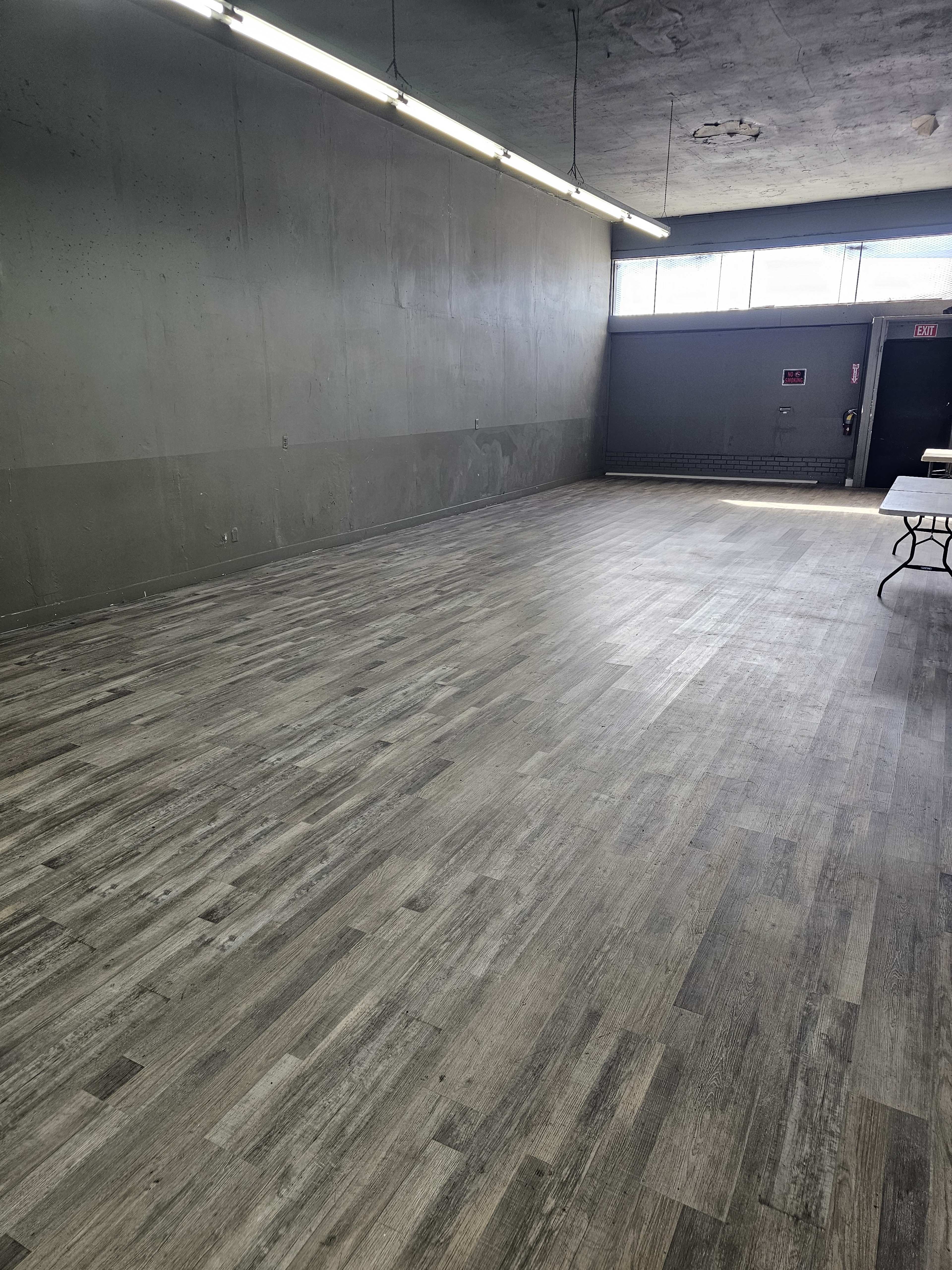 The image shows a spacious, empty room with a wooden floor and gray walls, featuring overhead lighting and a single table along one side.