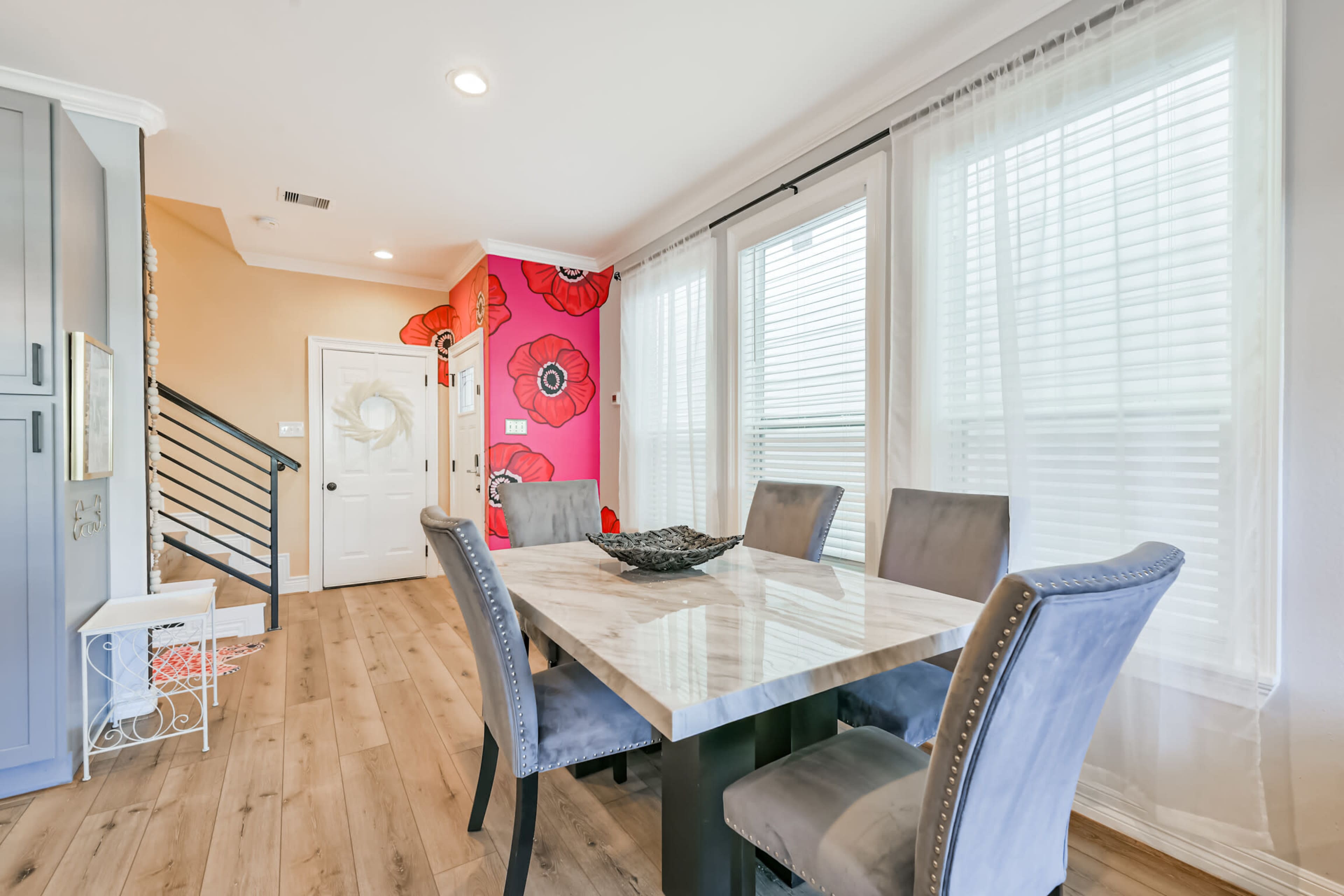 A dining area features a marble-top table surrounded by gray upholstered chairs, with large windows allowing natural light and colorful floral artwork on the wall.