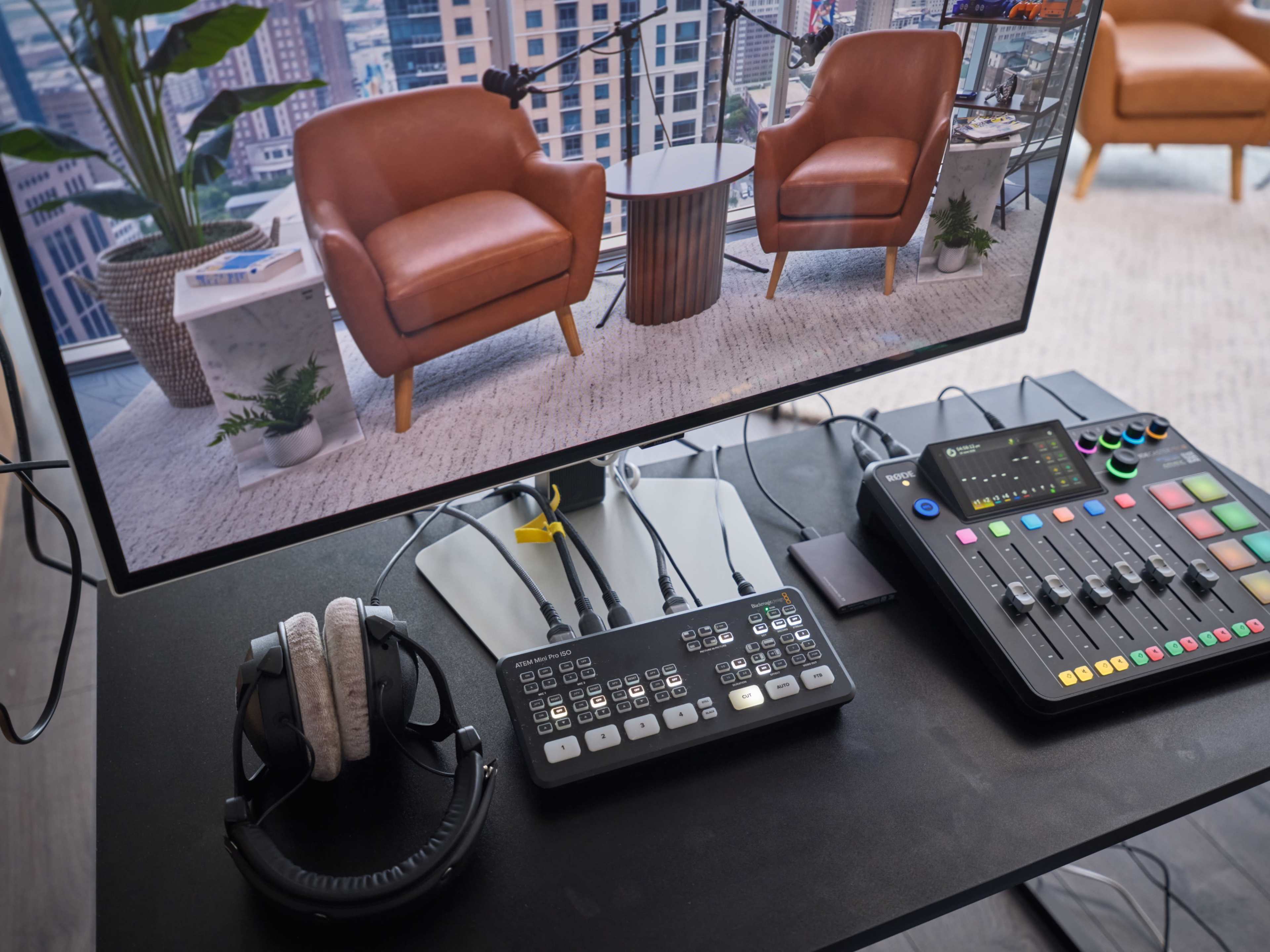 A monitor displays a virtual set with two brown chairs and a small table, while various audio and video equipment, including a control panel and headphones, are arranged on a desk.
