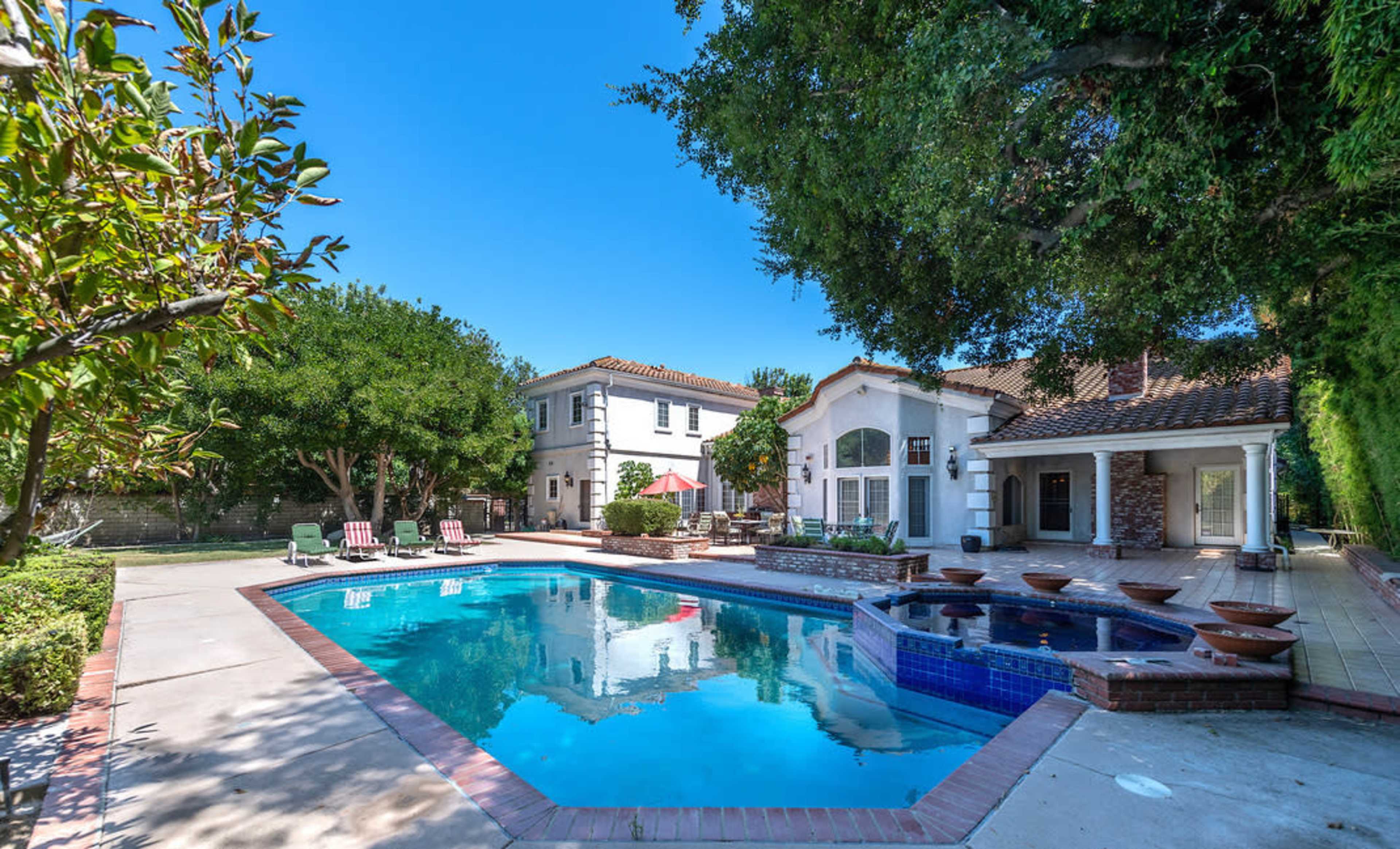 The image shows a landscaped backyard featuring a swimming pool, a hot tub, and an adjacent house with two distinct architectural styles.