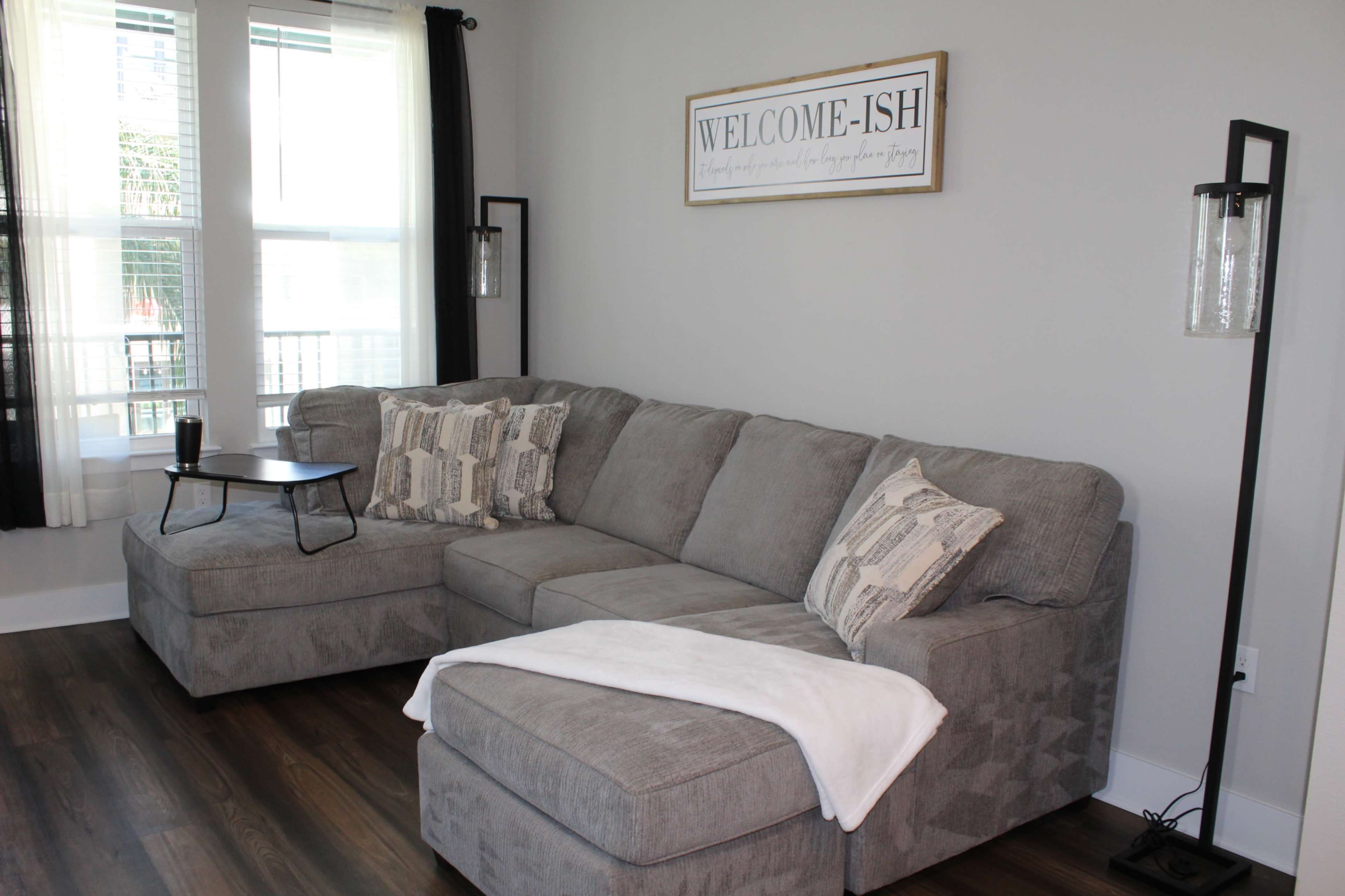 A gray sectional sofa with patterned pillows, a small side table, and a wall sign that reads "WELCOME-ISH" in a well-lit room with large windows.