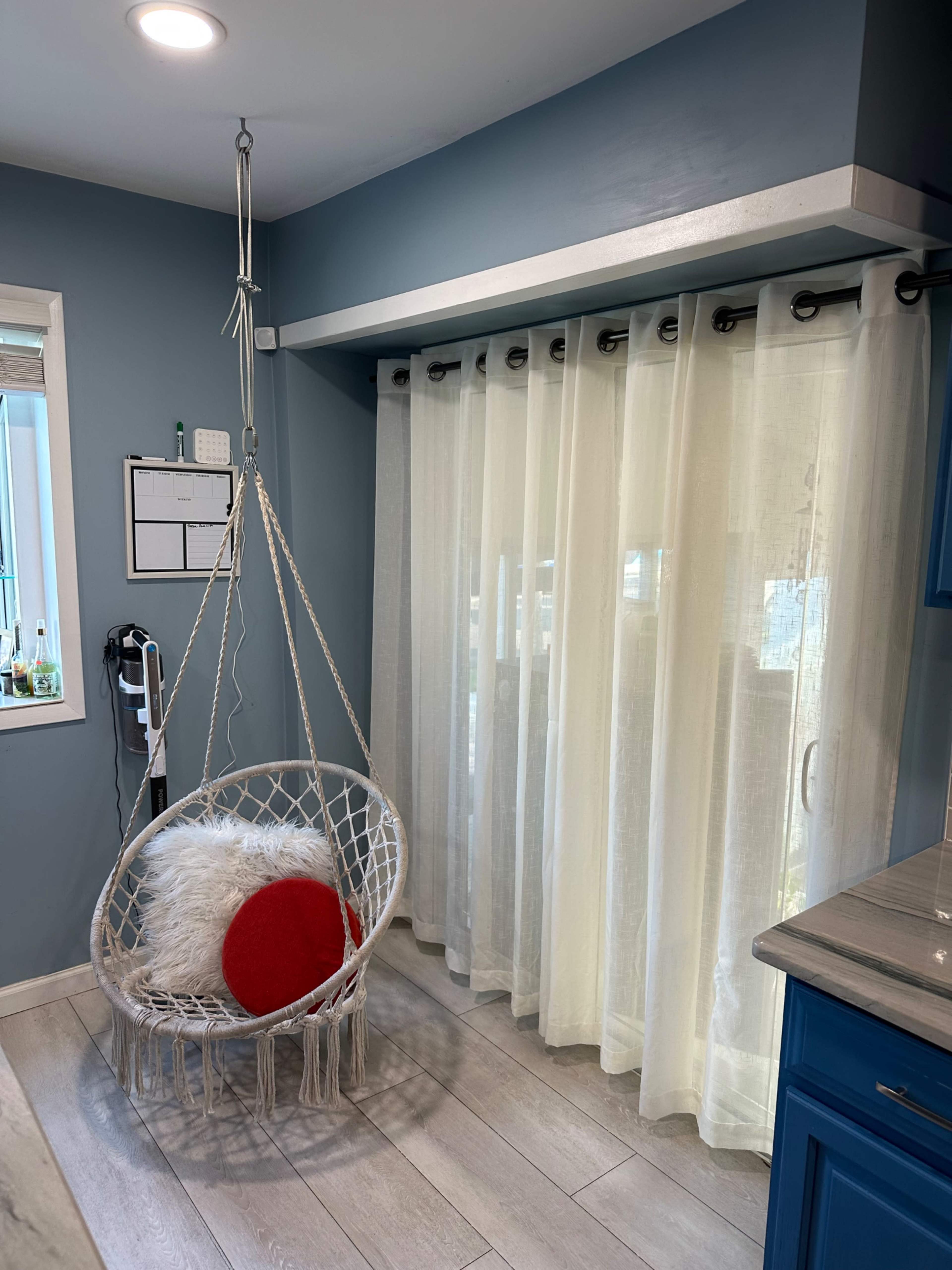 A hanging chair is positioned beside sheer white curtains in a blue-walled room, with a red cushion placed inside the chair.