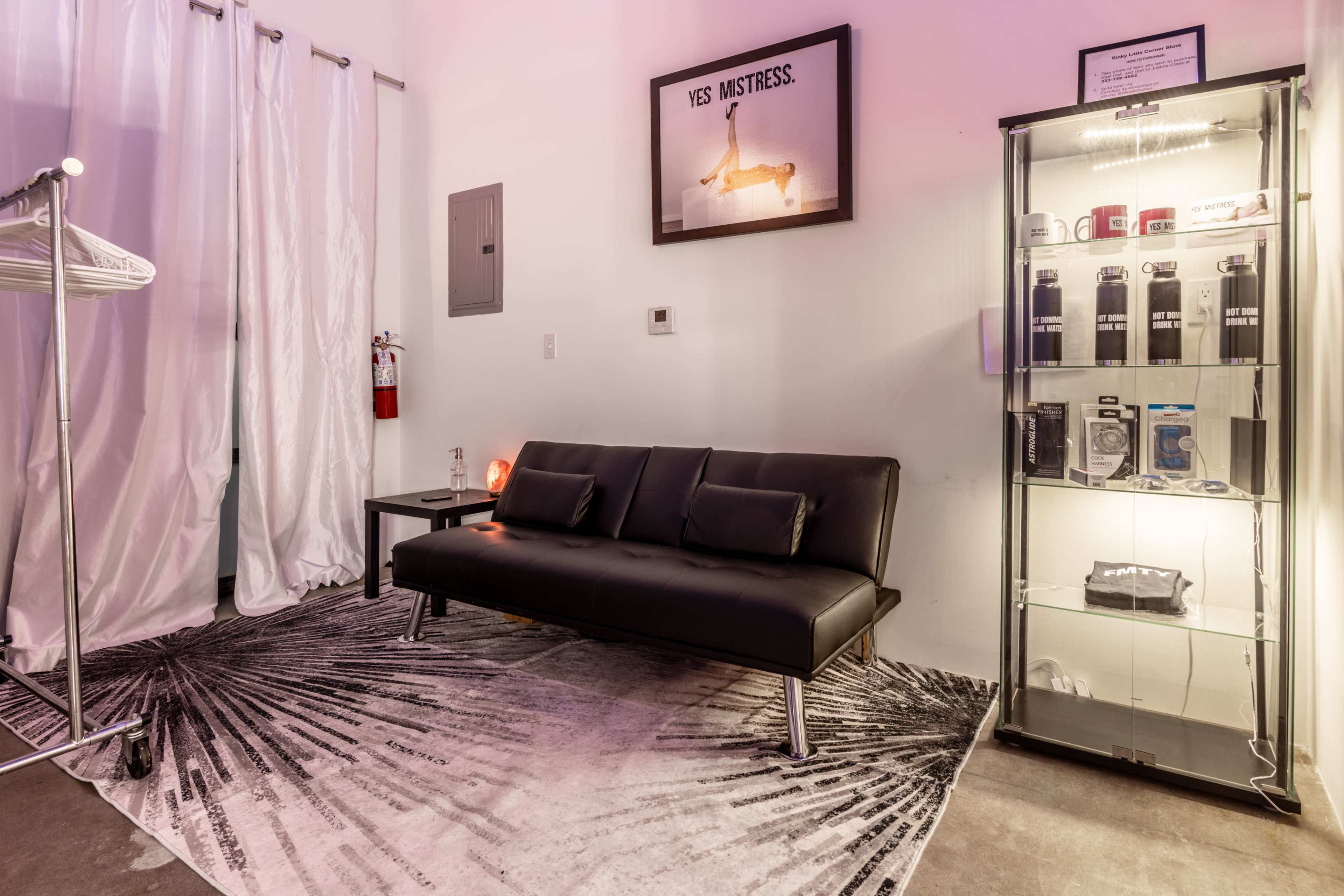 The image shows a minimalist interior space featuring a black couch, a glass display case with various items, and white curtains, all against a backdrop of a framed poster.