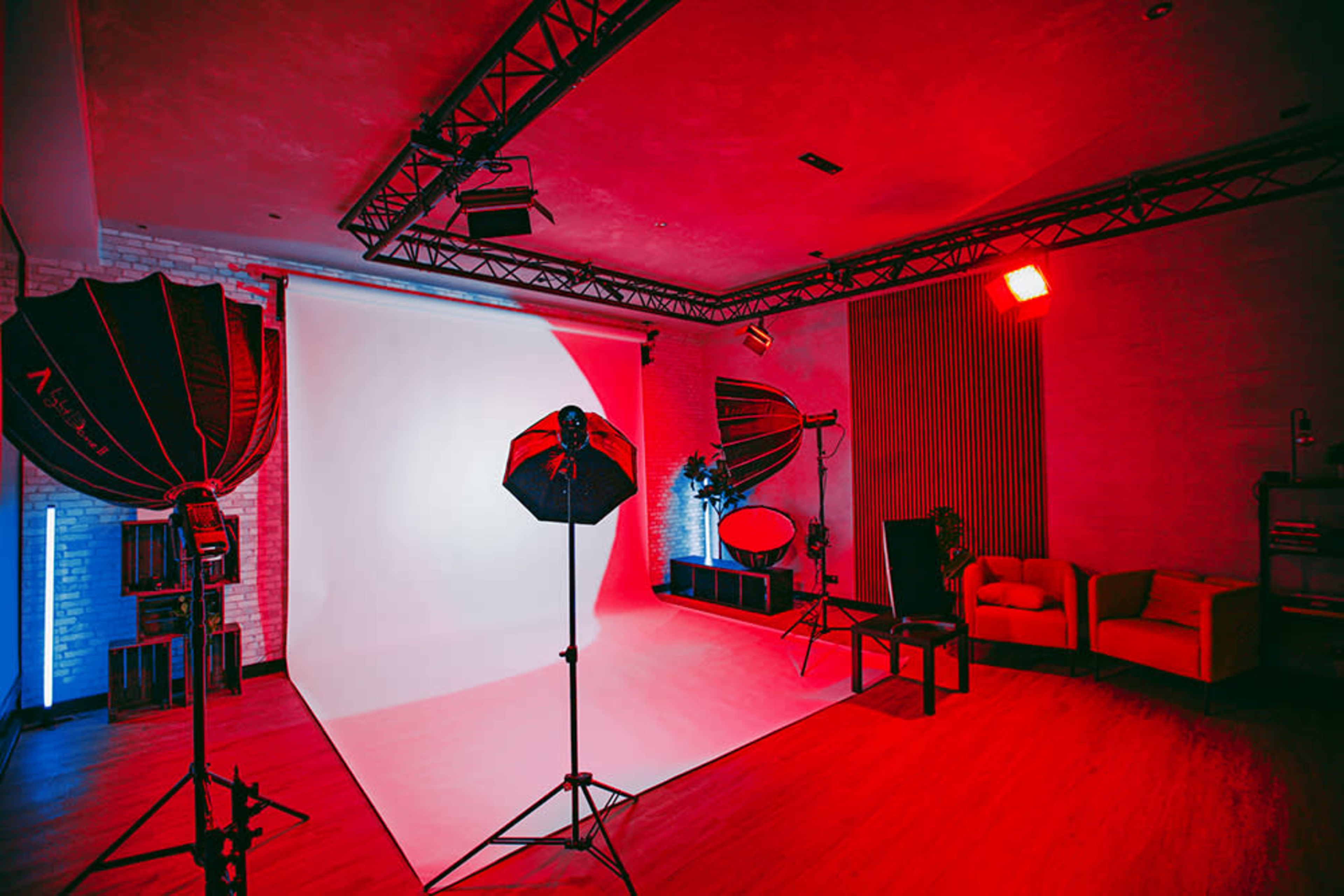 The image shows a photography studio with red lighting, equipped with softboxes, a white backdrop, and a seating area.