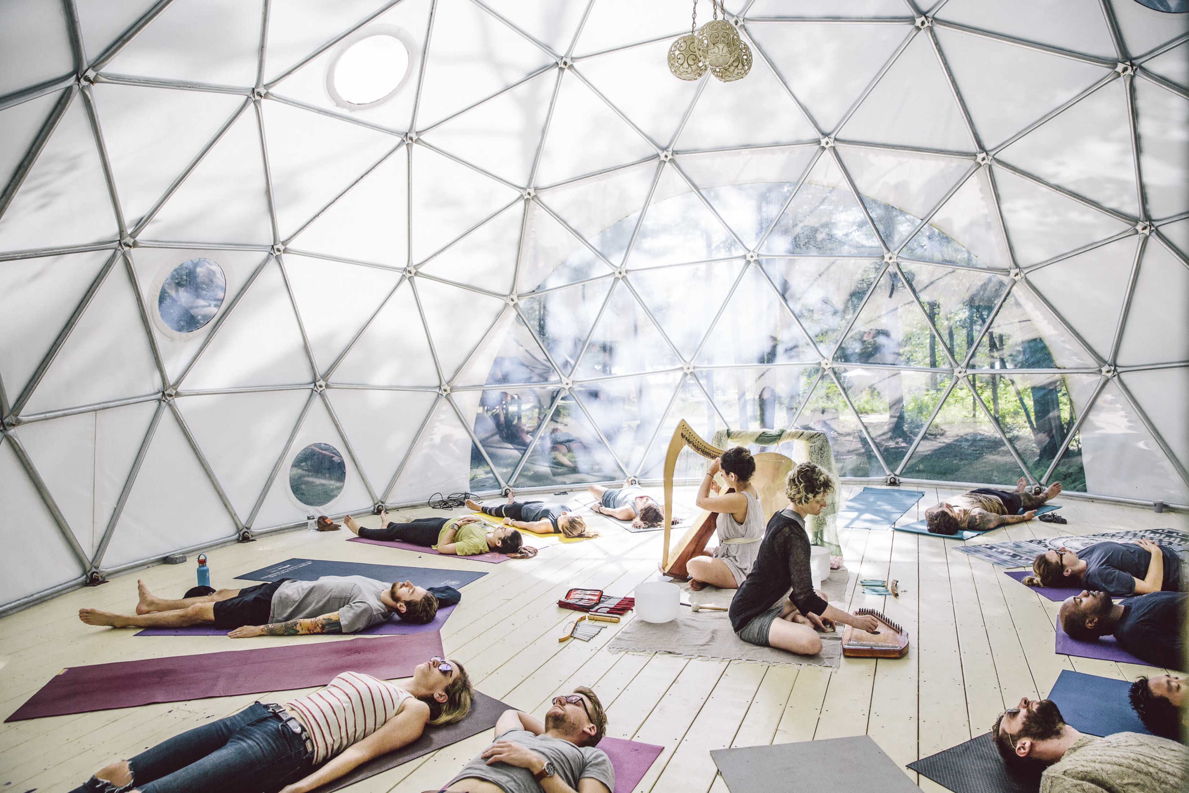 A group of people lies on yoga mats inside a geodesic dome, while a musician plays a harp in the center.