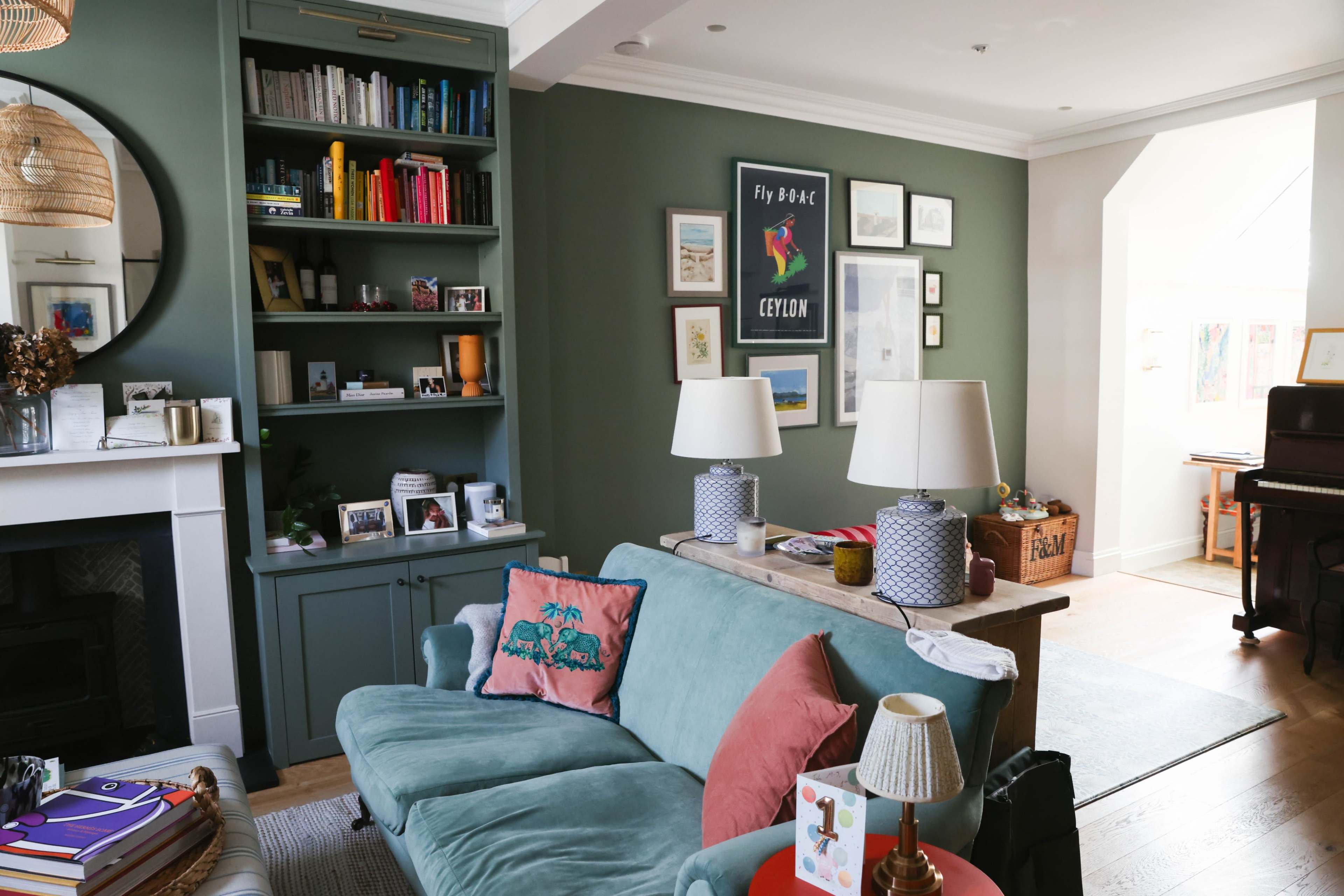 The image shows a cozy living room with a teal sofa, shelves filled with books and framed pictures, and a piano in an adjoining space.
