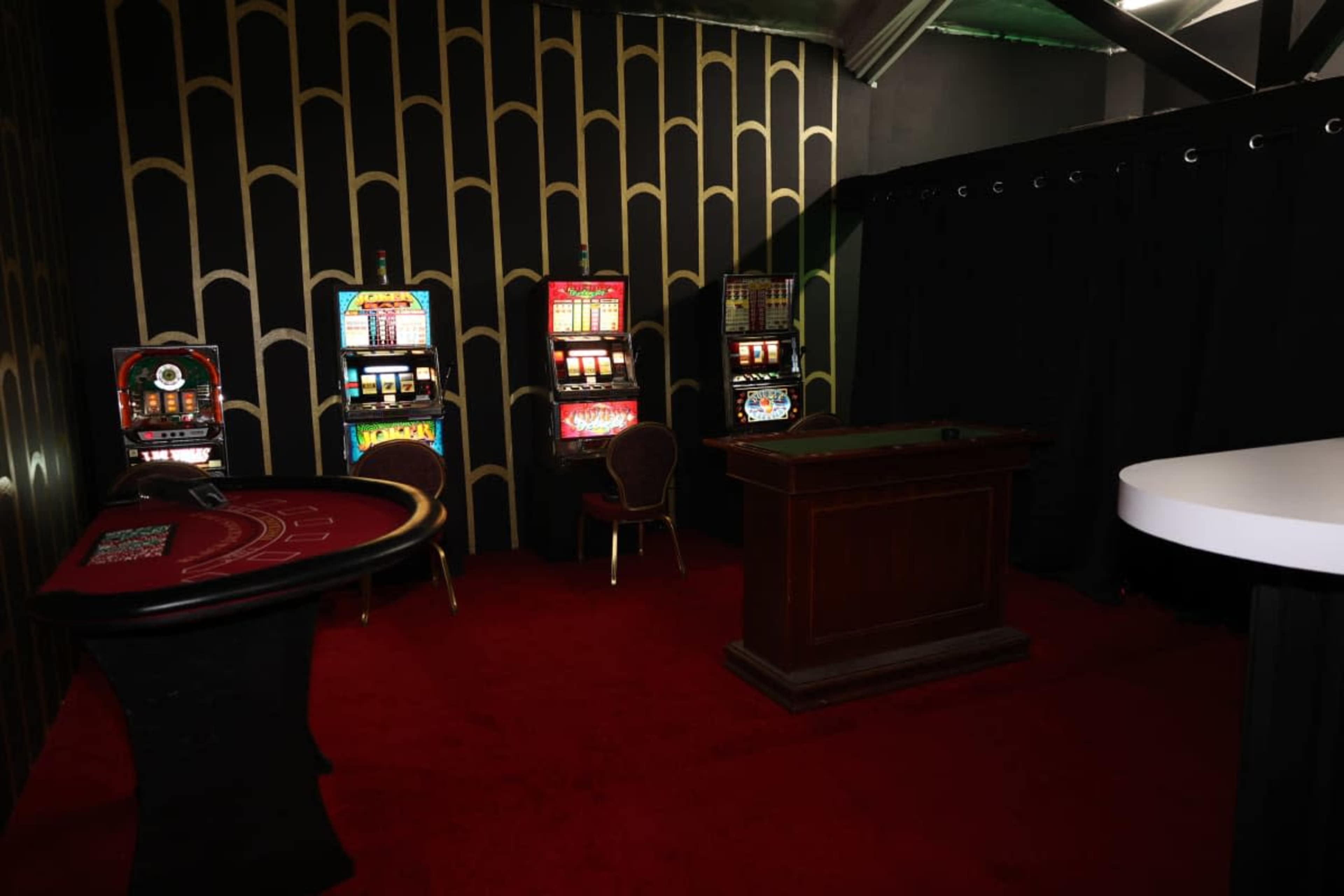 The image shows a small gaming area featuring a roulette table, a wooden dealer's table, and several slot machines against a patterned black and gold wall.