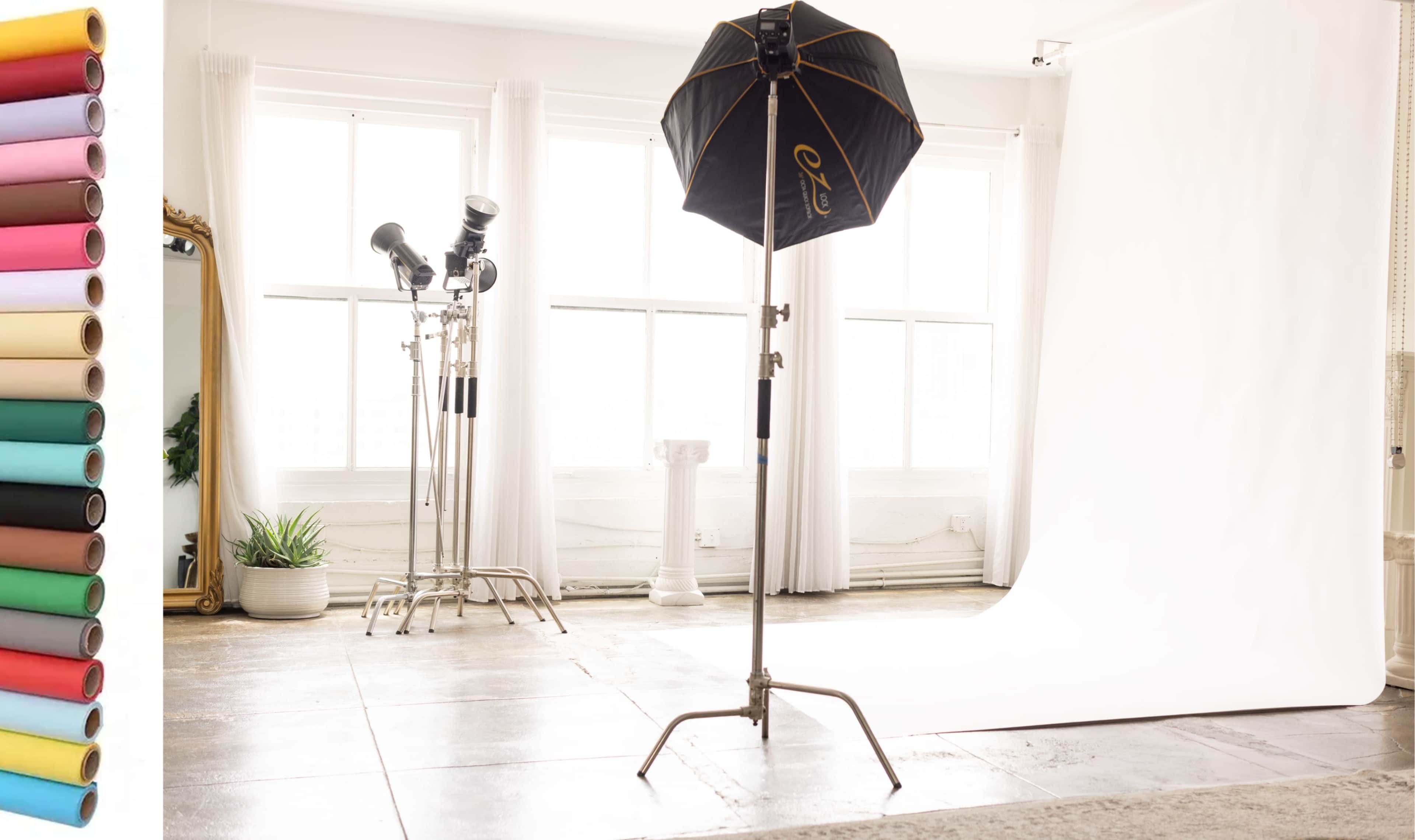 A photography studio features a backdrop with a stand, lighting equipment, and a variety of colored paper rolls on one side.
