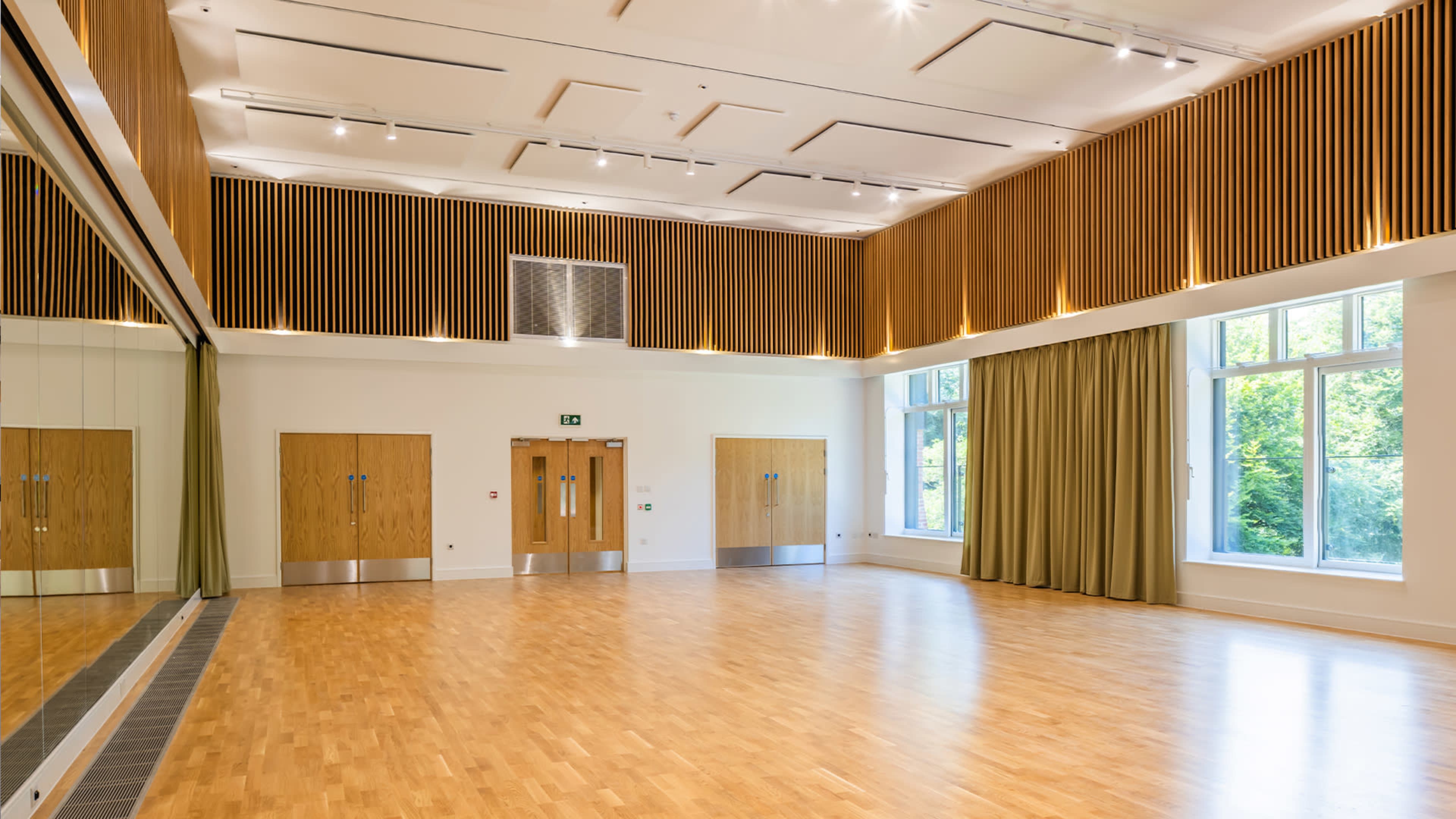 A spacious room with wooden flooring, large windows, and a wall adorned with wooden slats, along with two sets of double doors.