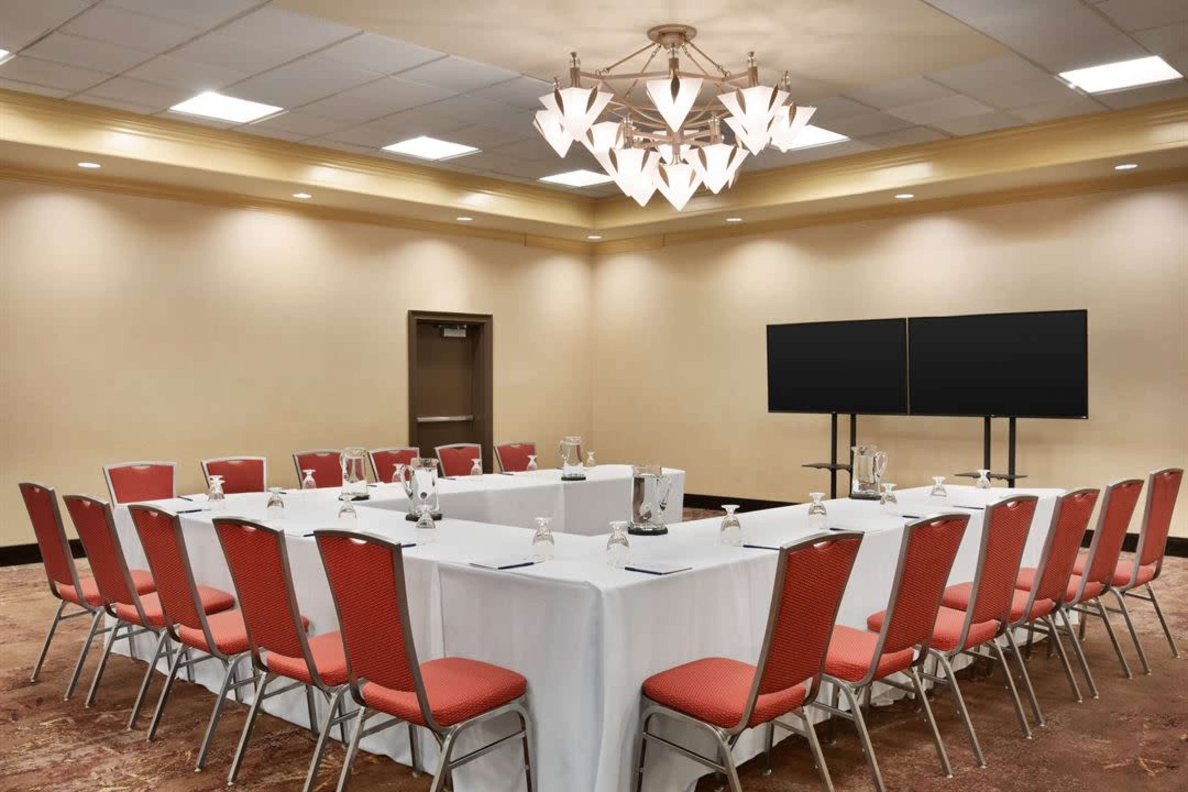 The image shows a conference room set up with a U-shaped arrangement of tables covered in white cloth and surrounded by red chairs, with two screens at one end.