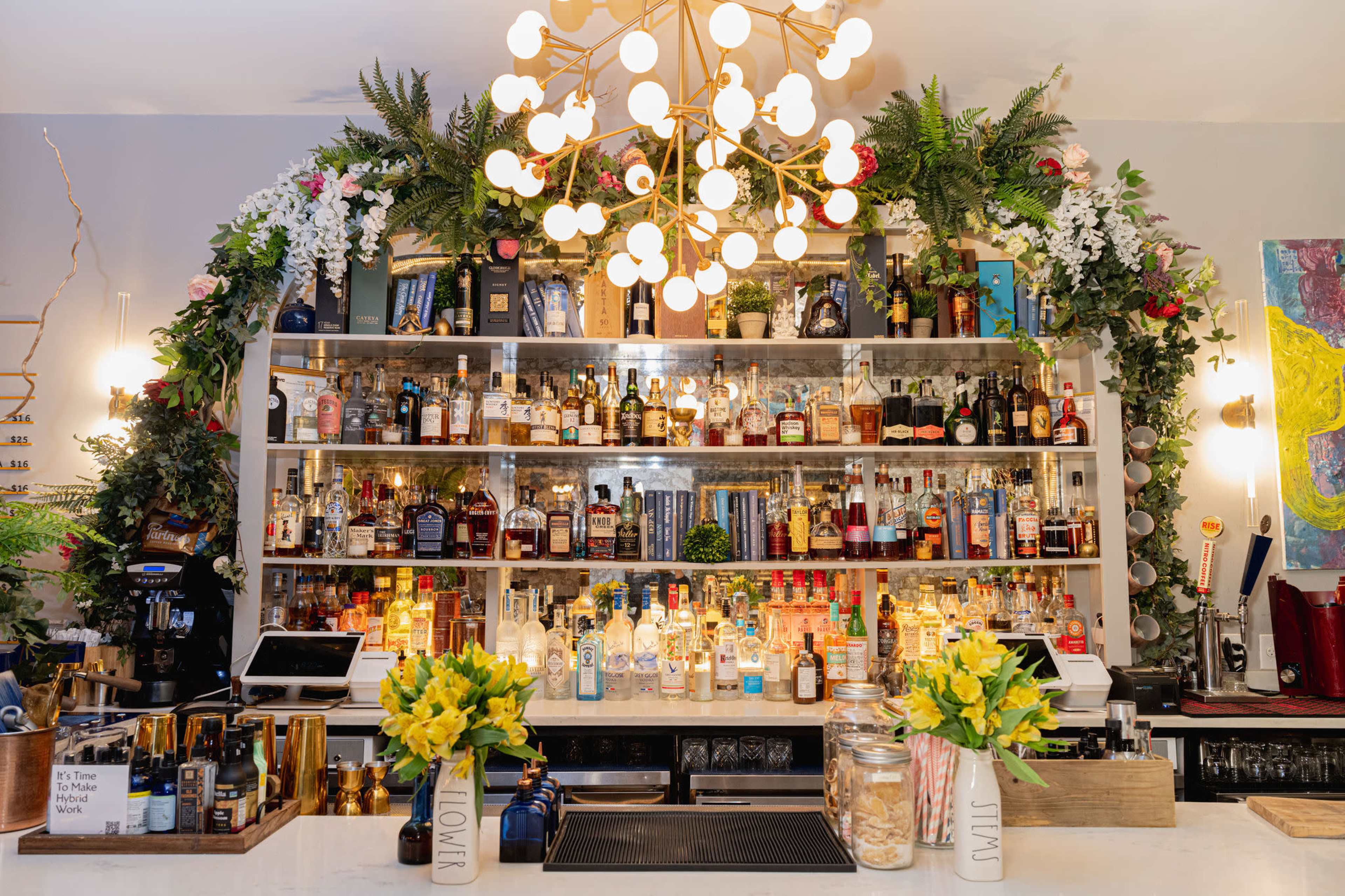A well-organized bar counter with various bottles of liquor displayed on shelves adorned with greenery and floral arrangements, beneath a modern chandelier.