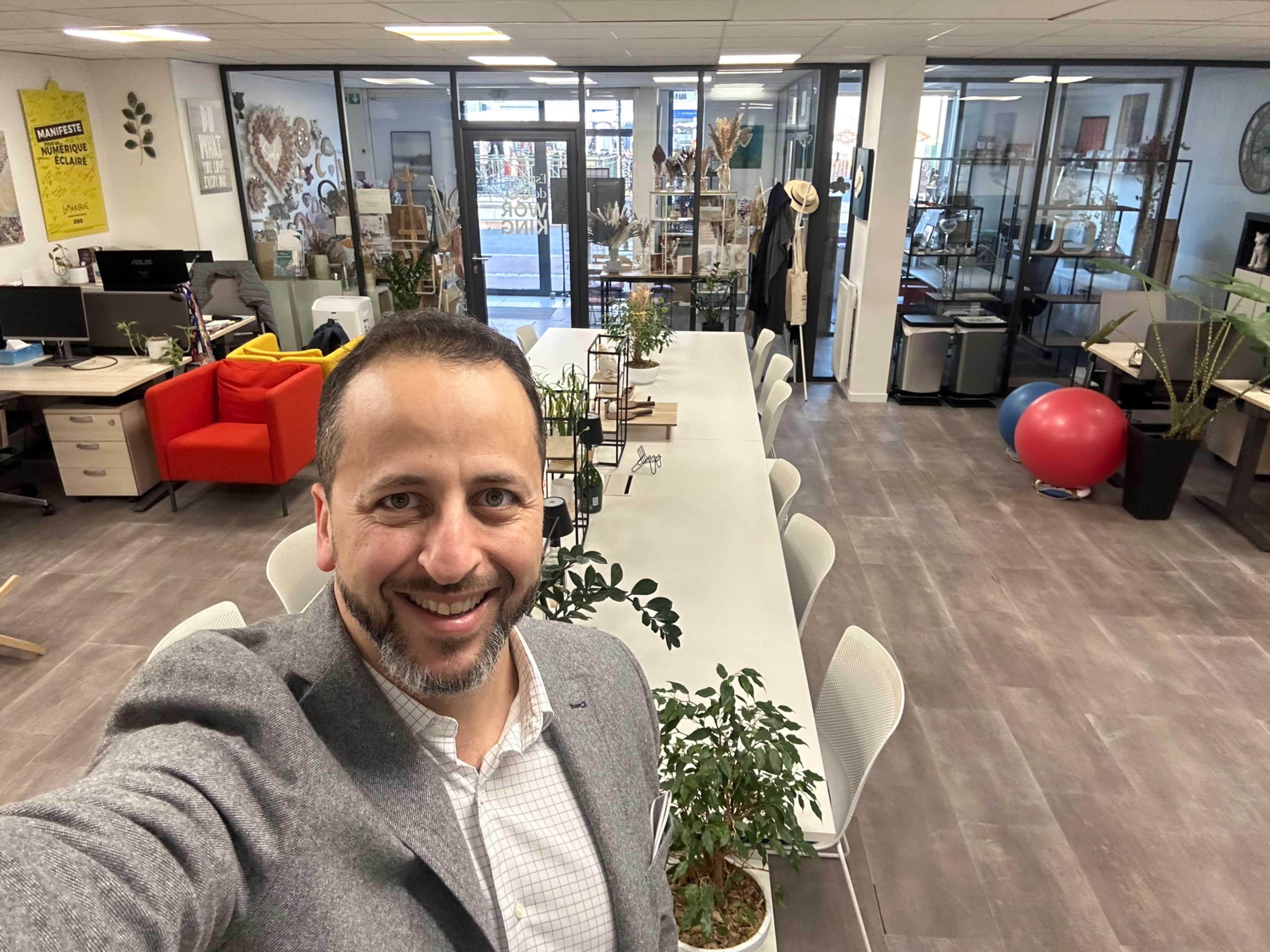 A man stands in an office space featuring a long white table, plants, and various workstations with colorful furniture.