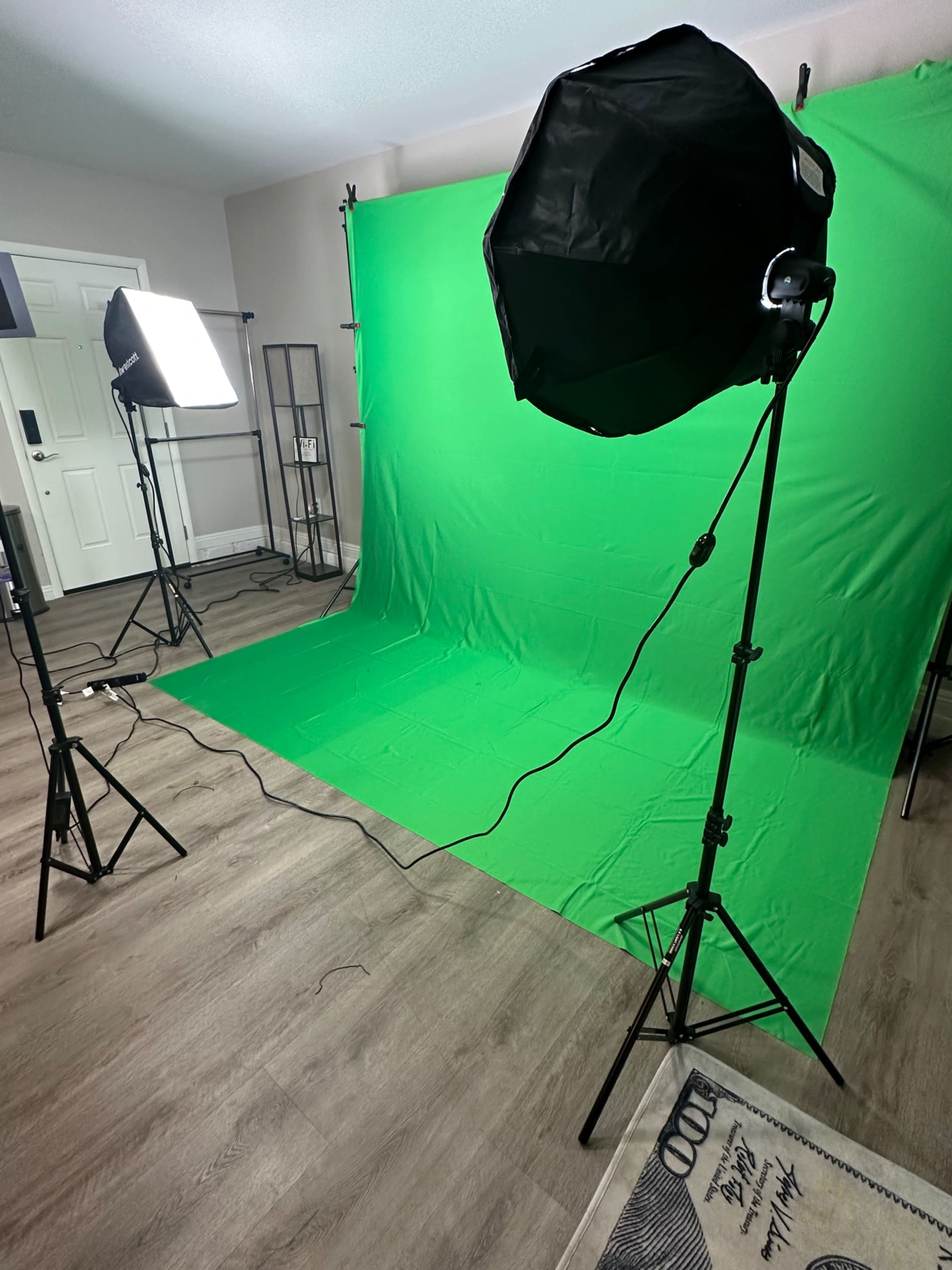 A studio setup featuring a green screen, two lighting equipment stands, and wooden flooring.