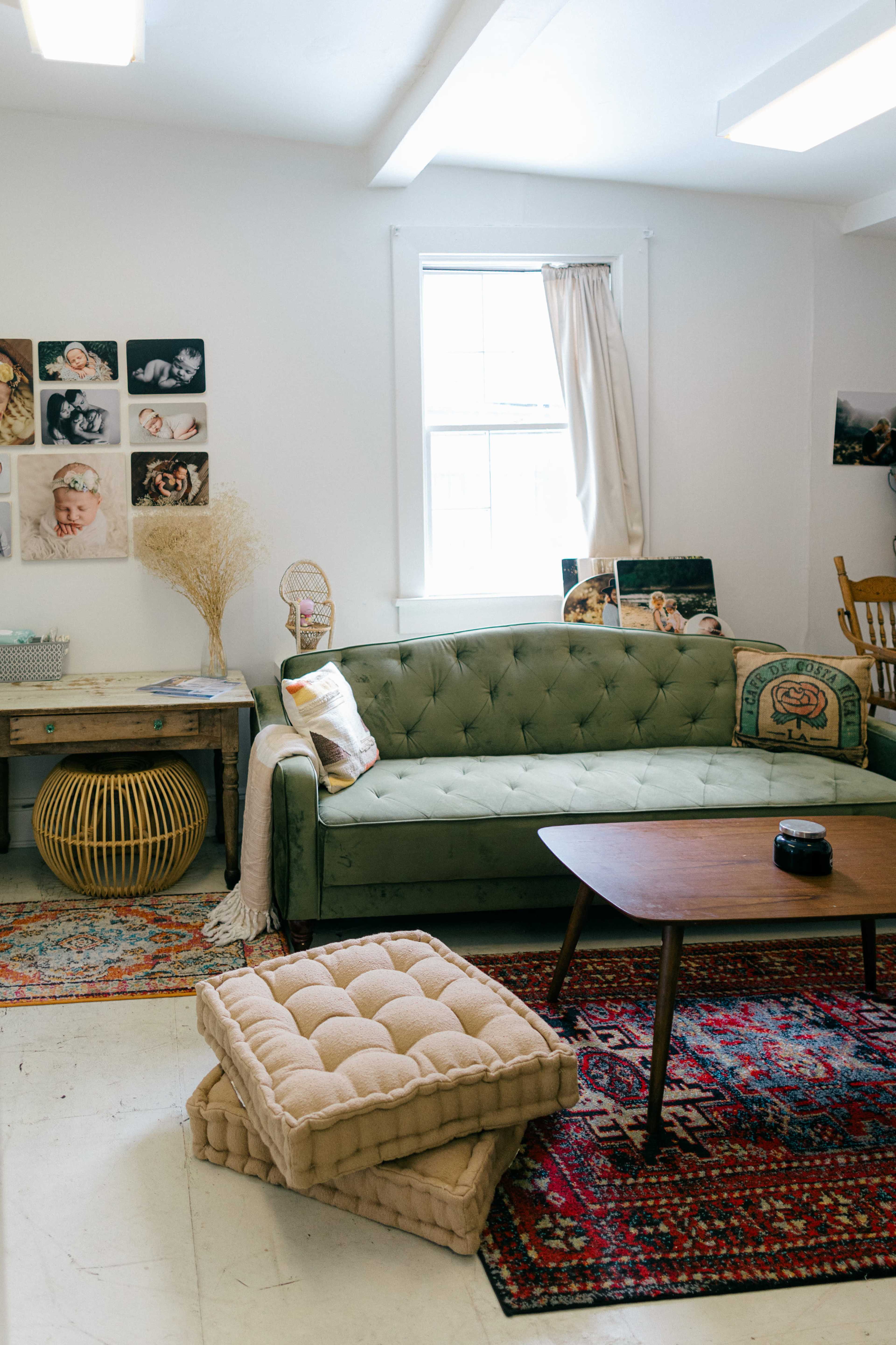 The image shows a cozy room featuring a green sofa, a wooden table, decorative pillows, and art on the walls, with a patterned rug on the floor.